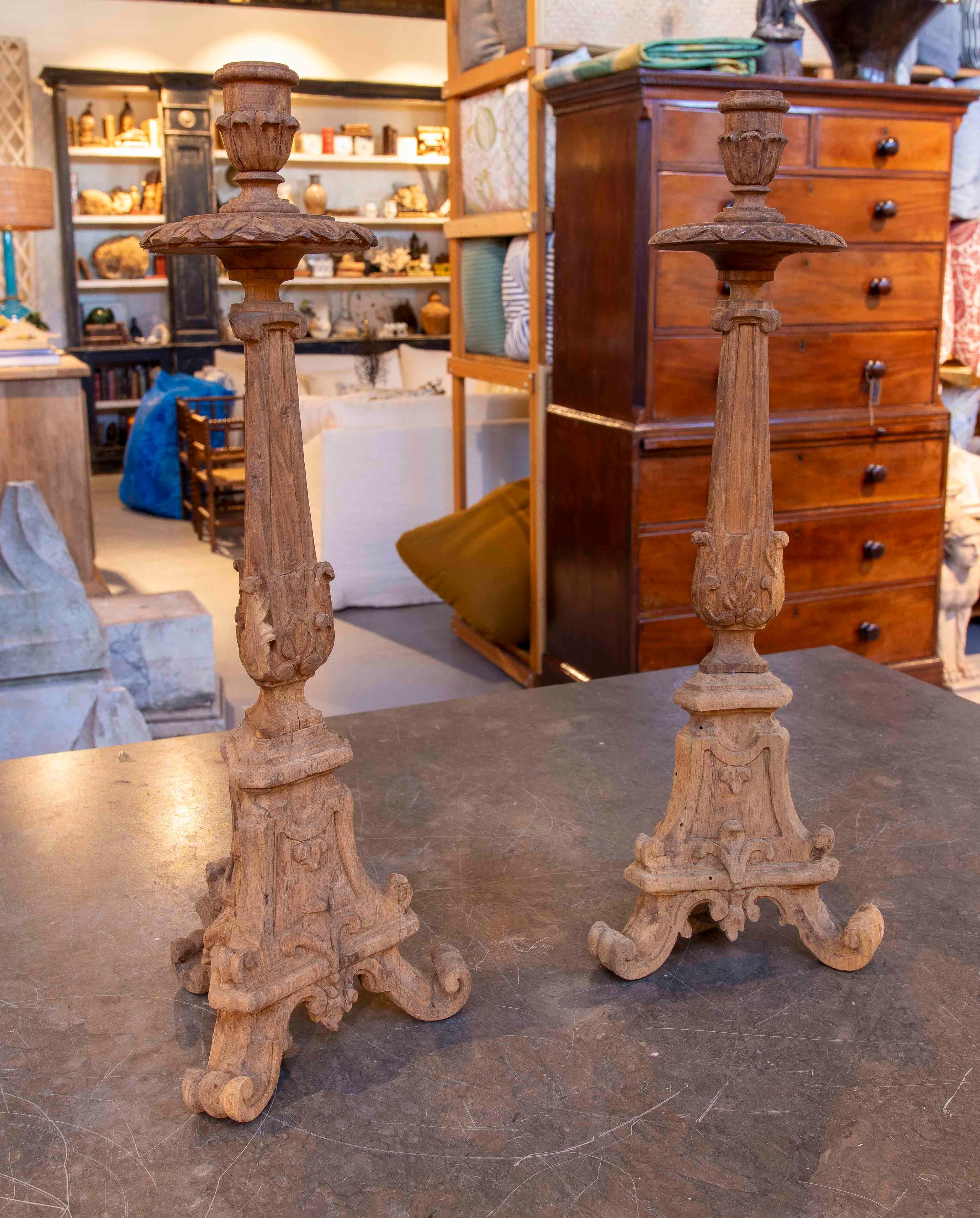 An elegant pair of carved wooden baroque candlesticks featuring a striking tripod base of curved scrolls and embossed floral motifs. The delicately raised centerpiece is decorated with classic carvings reminiscent of acanthus leaves and symmetrical