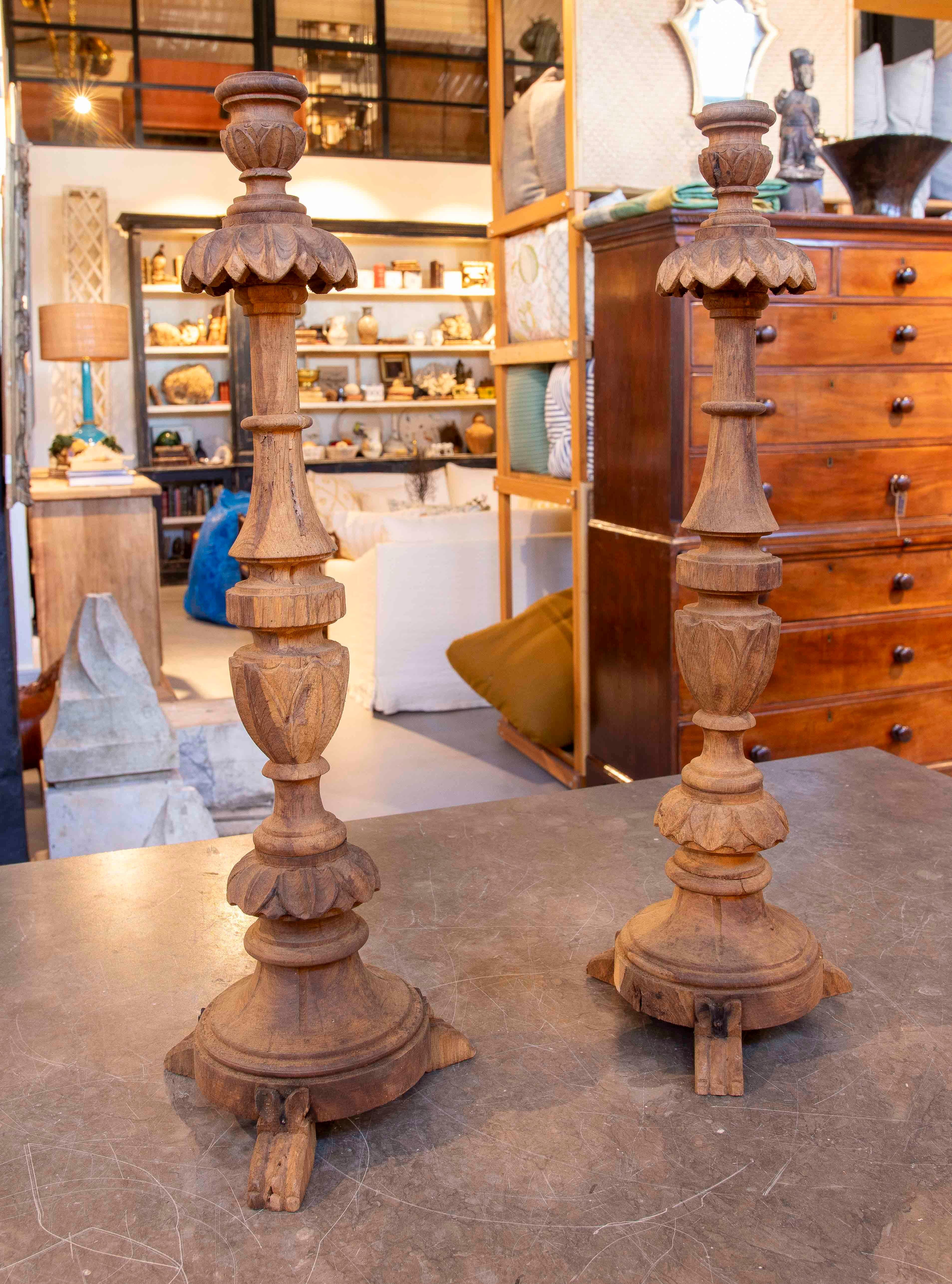 An elegant pair of tall candlesticks crafted from carved wood with a natural, unvarnished finish, highlighting the material's elegance and texture. The design features a sturdy, three-legged, claw-shaped base, a stem decorated with plant motifs, and