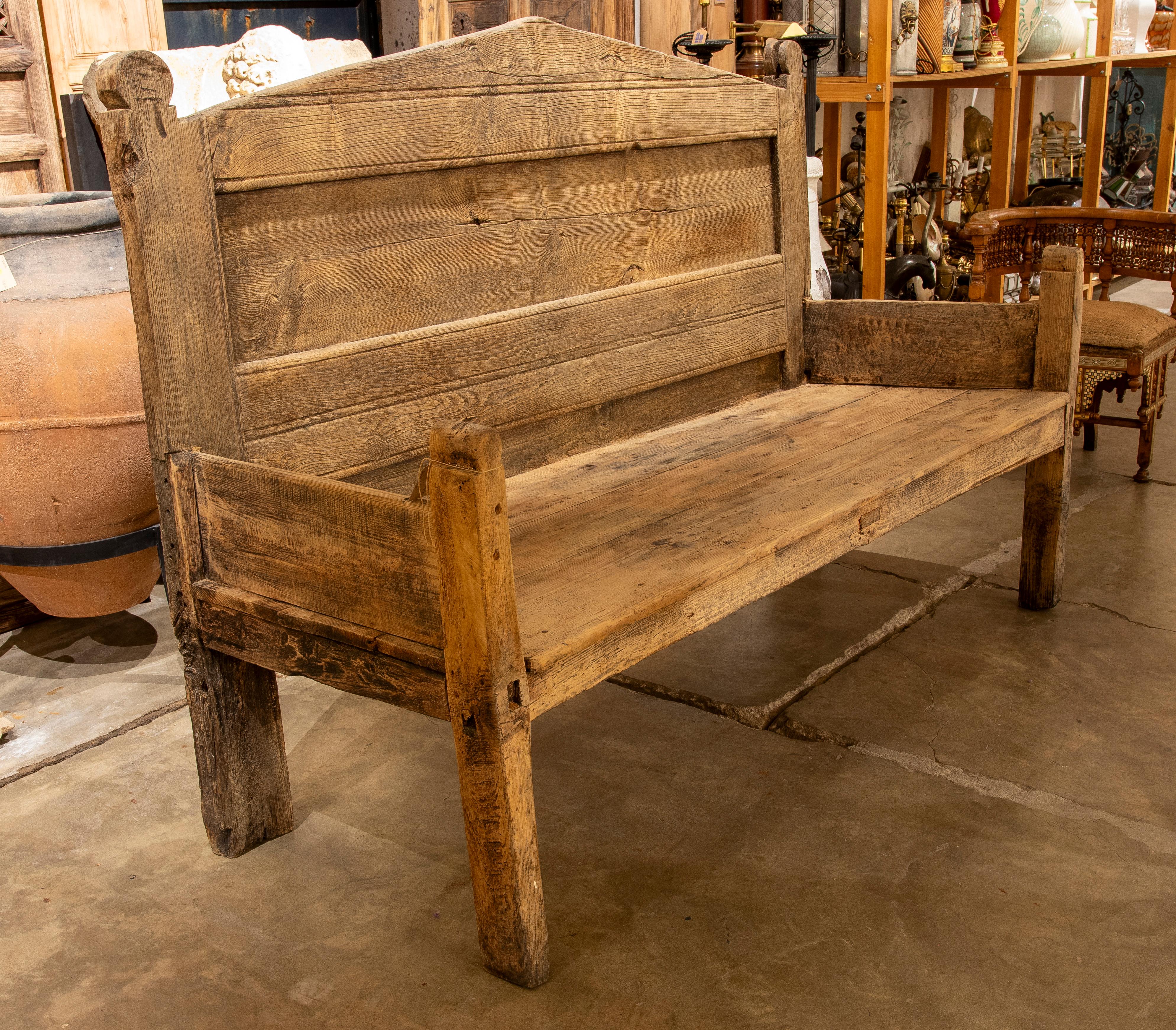 Este robusto banco está hecho de madera maciza envejecida, con un diseño auténtico y sin pretensiones que resalta su carácter rústico y artesanal. El respaldo alto, compuesto por tablones horizontales, y los reposabrazos rectos le dan una presencia