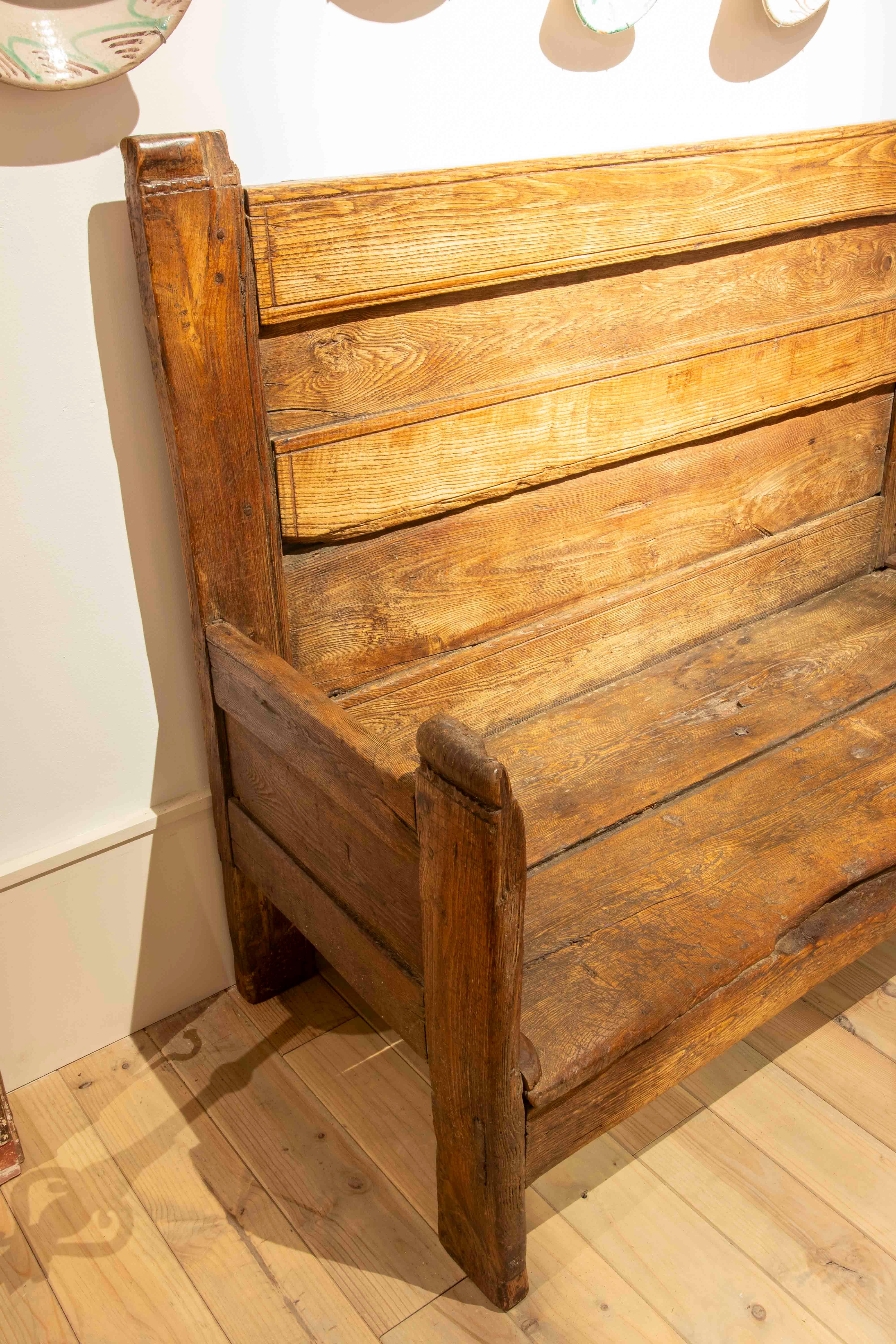 Un banco antiguo de madera maciza con acabado natural, que conserva su pátina original y los signos del tiempo que reflejan su autenticidad. De líneas sencillas y robustas, este banco presenta un respaldo alto compuesto por tablones verticales,