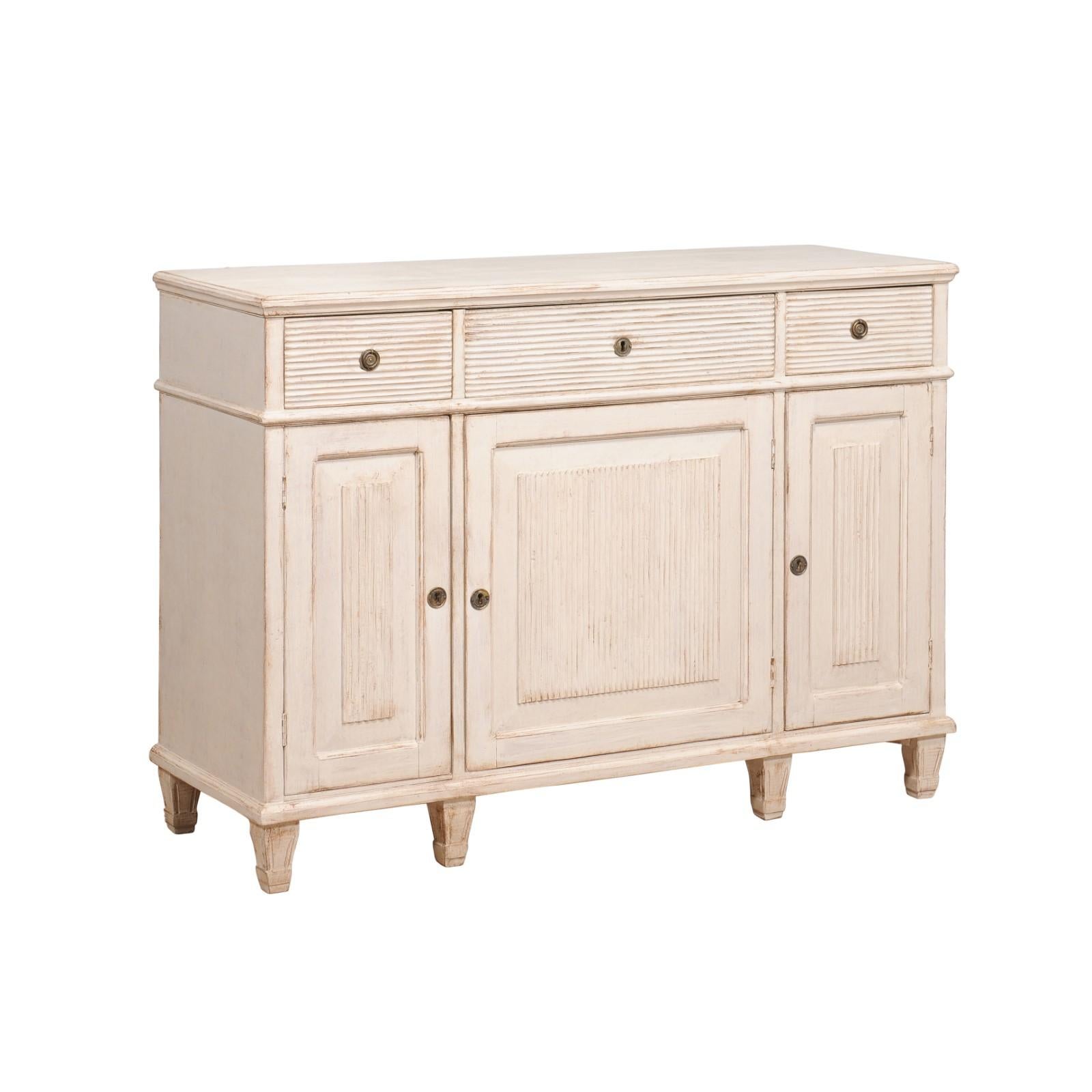 A Swedish 1850s sideboard from Dalarna, with off white, antique beige painted finish, three drawers over three doors, carved reeded motifs and short tapering feet. Bask in the elegant allure of this 1850s Swedish Sideboard, a charming artifact from