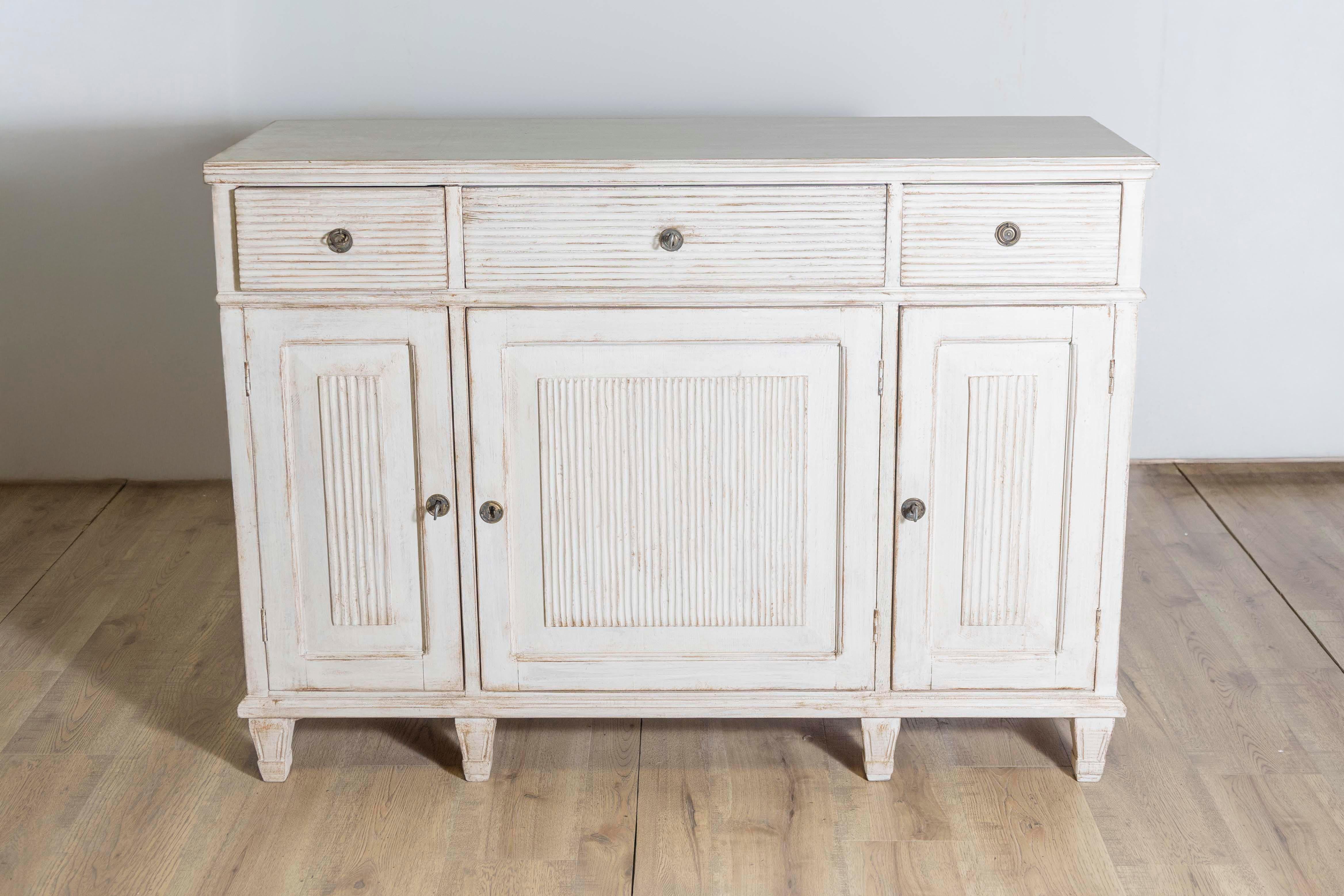A Swedish 1850s sideboard from Dalarna, with off white, antique beige painted finish, three drawers over three doors, carved reeded motifs and short tapering feet. Bask in the elegant allure of this 1850s Swedish Sideboard, a charming artifact from
