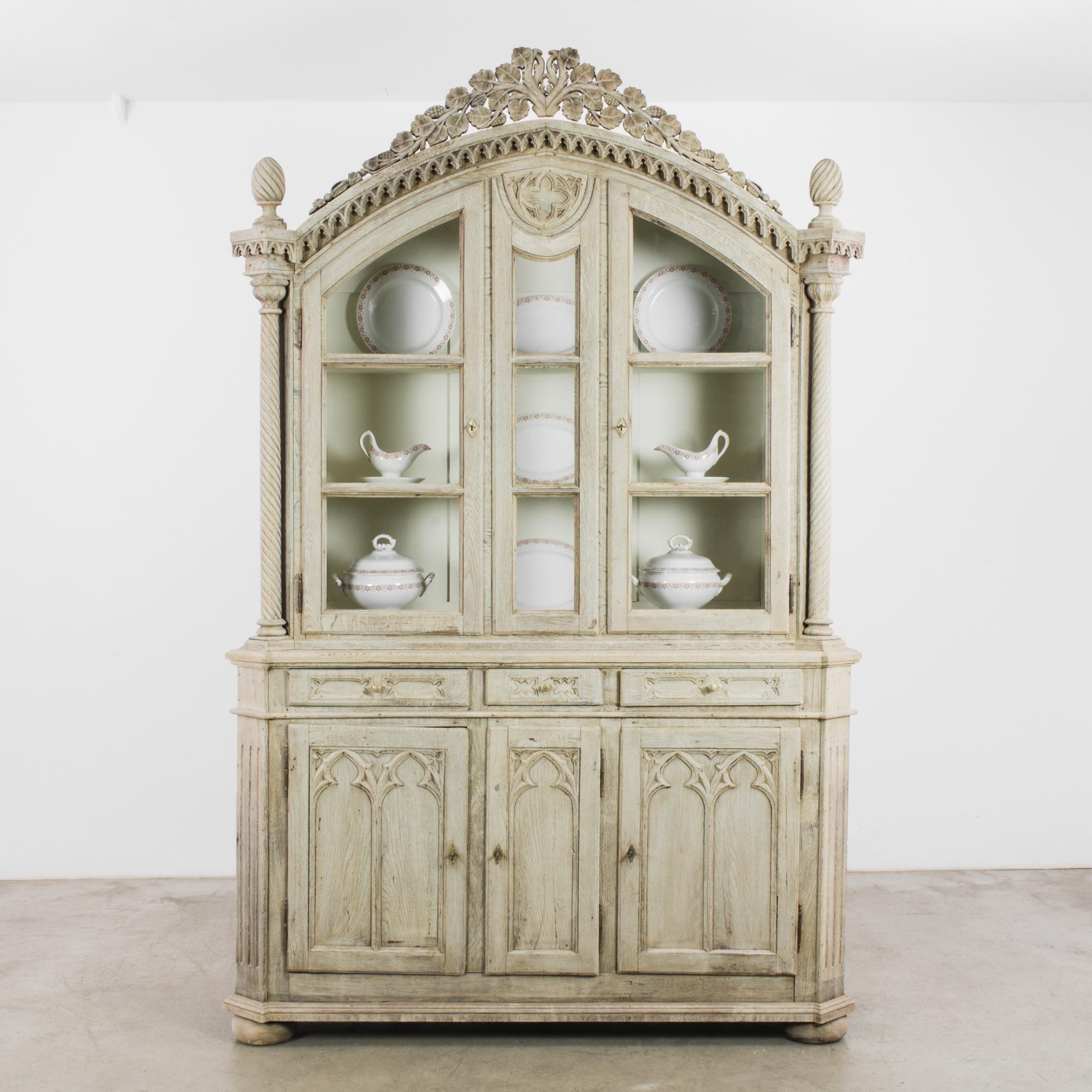An antique oak vitrine circa 1860, sourced from Belgium. A gothic silhouette is festooned with elaborate ornament: Gothic dorr panels, spiral columns, a quatrefoil centerpiece. The oak has been restored to a pale, moonlit tone, a natural finish