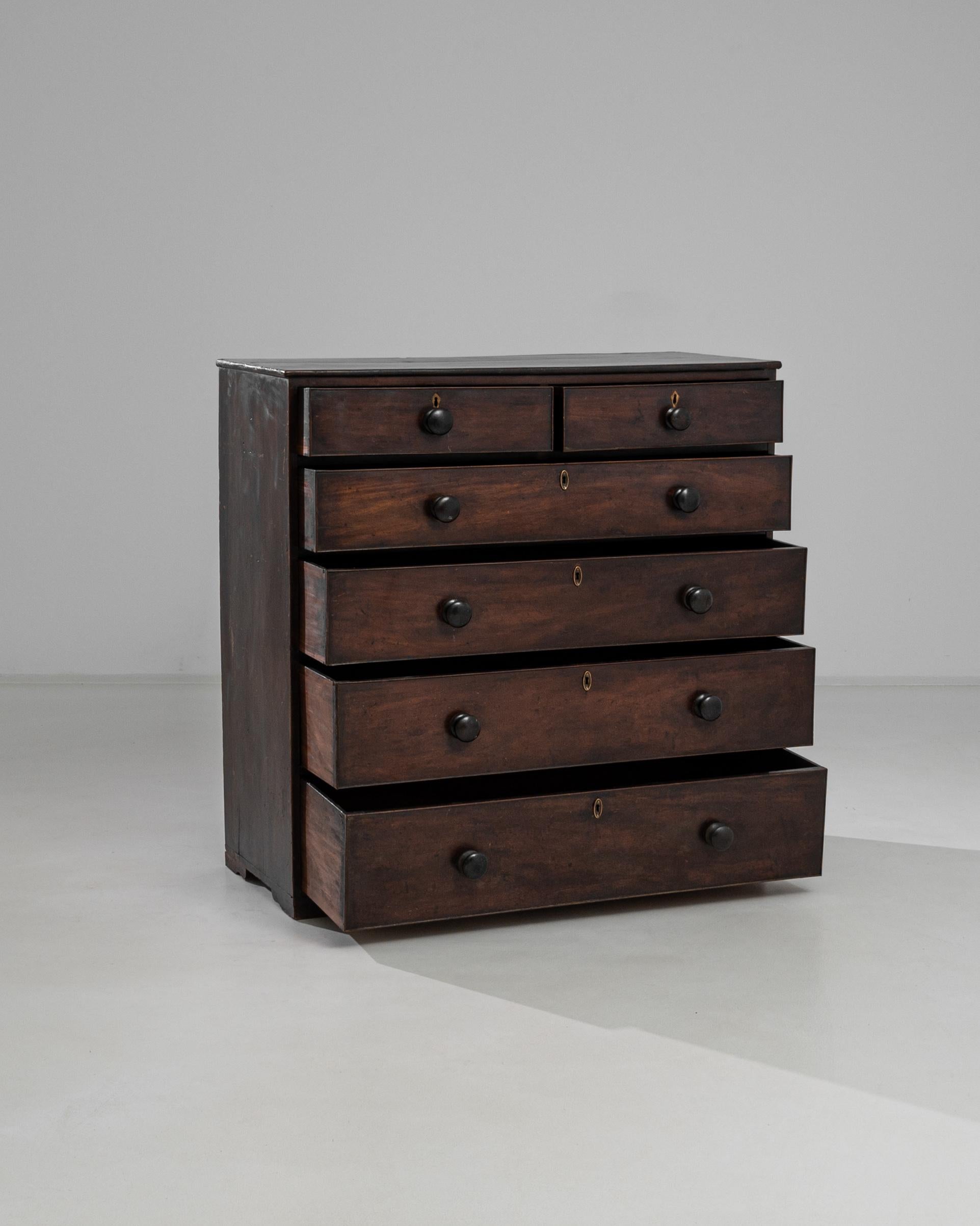 This 1860s British Wooden Chest of Drawers is a rich tapestry of history and craftsmanship. With its original patina, this piece carries the whispers of the past into the modern home. The well-worn wood tells a story of enduring strength, while the
