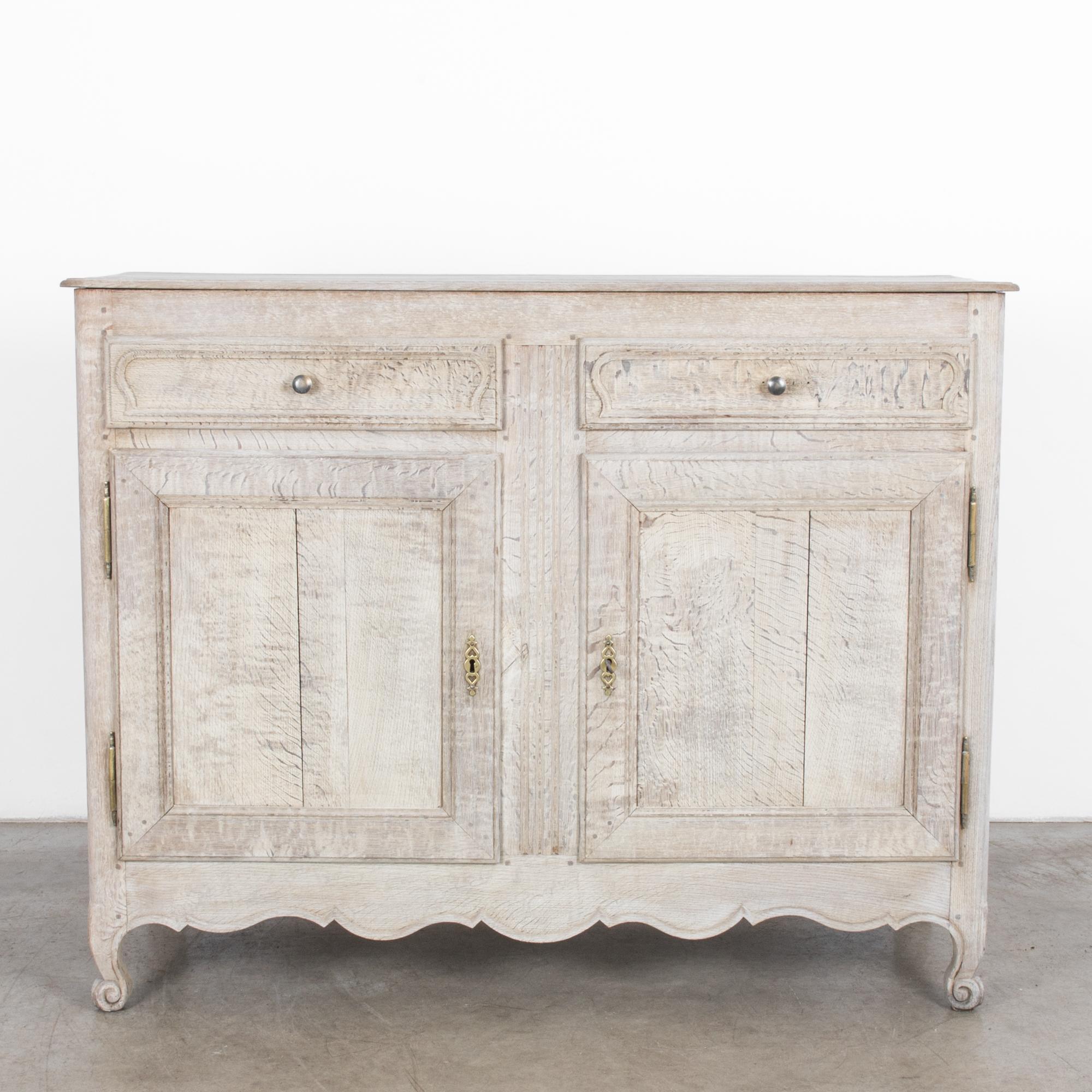 A bleached oak buffet from France, circa 1860. Two paneled doors open onto an interior shelf, above, twin drawers pull out with silver knobs. The curved paneling on the drawers echoes the elegant billows of the carved apron. Dainty scrolled feet