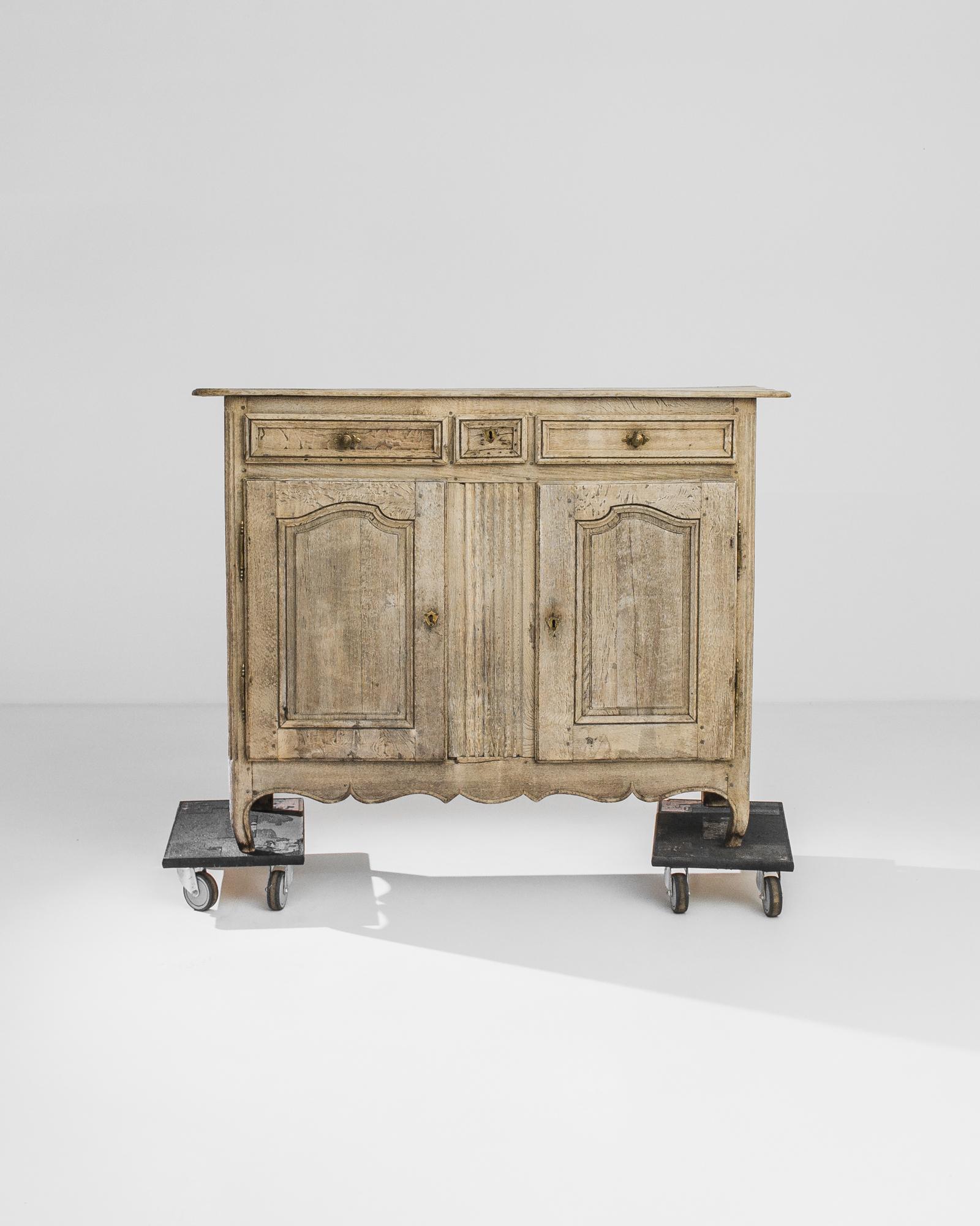 An oak buffet from 1860s France. Cabriole feet and a scalloped apron give an air of delicacy to the upright frame; original gilded drawer handles and a crest-shaped lock piece add a noble touch. The warm biscuit color of the restored wood lends a
