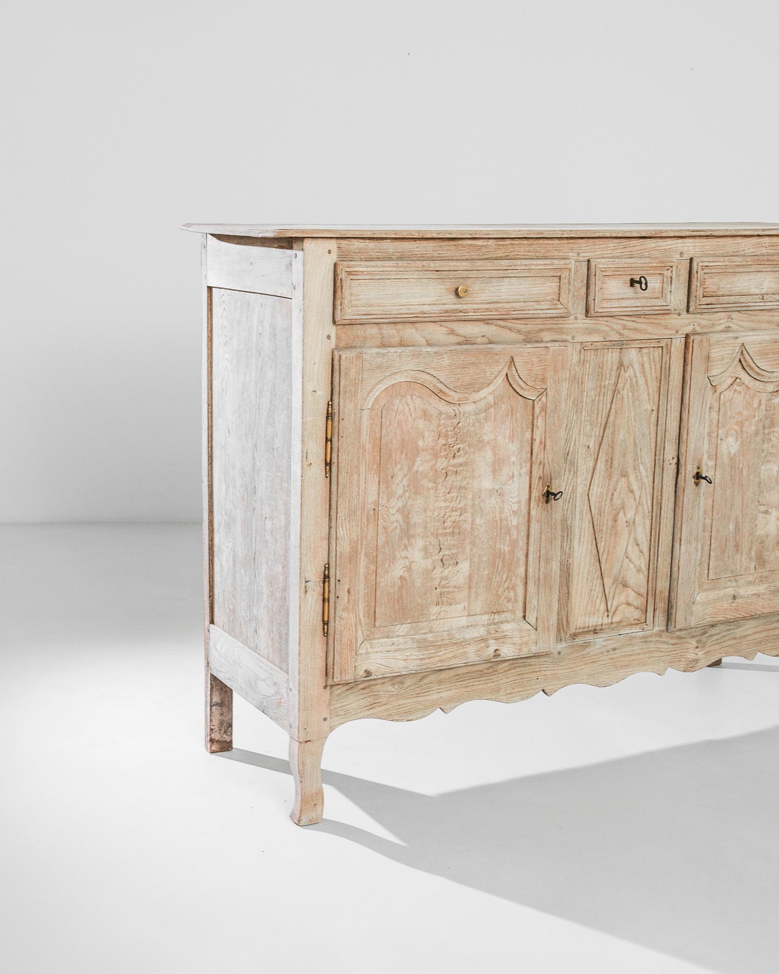 An oak buffet from 1860s France. The delicate finish of the restored wood, flushed with pale rose pink notes, accentuates the fluid curves and peaks of paneling and apron. A diamond-shaped motif on the central panel is mirrored in the original