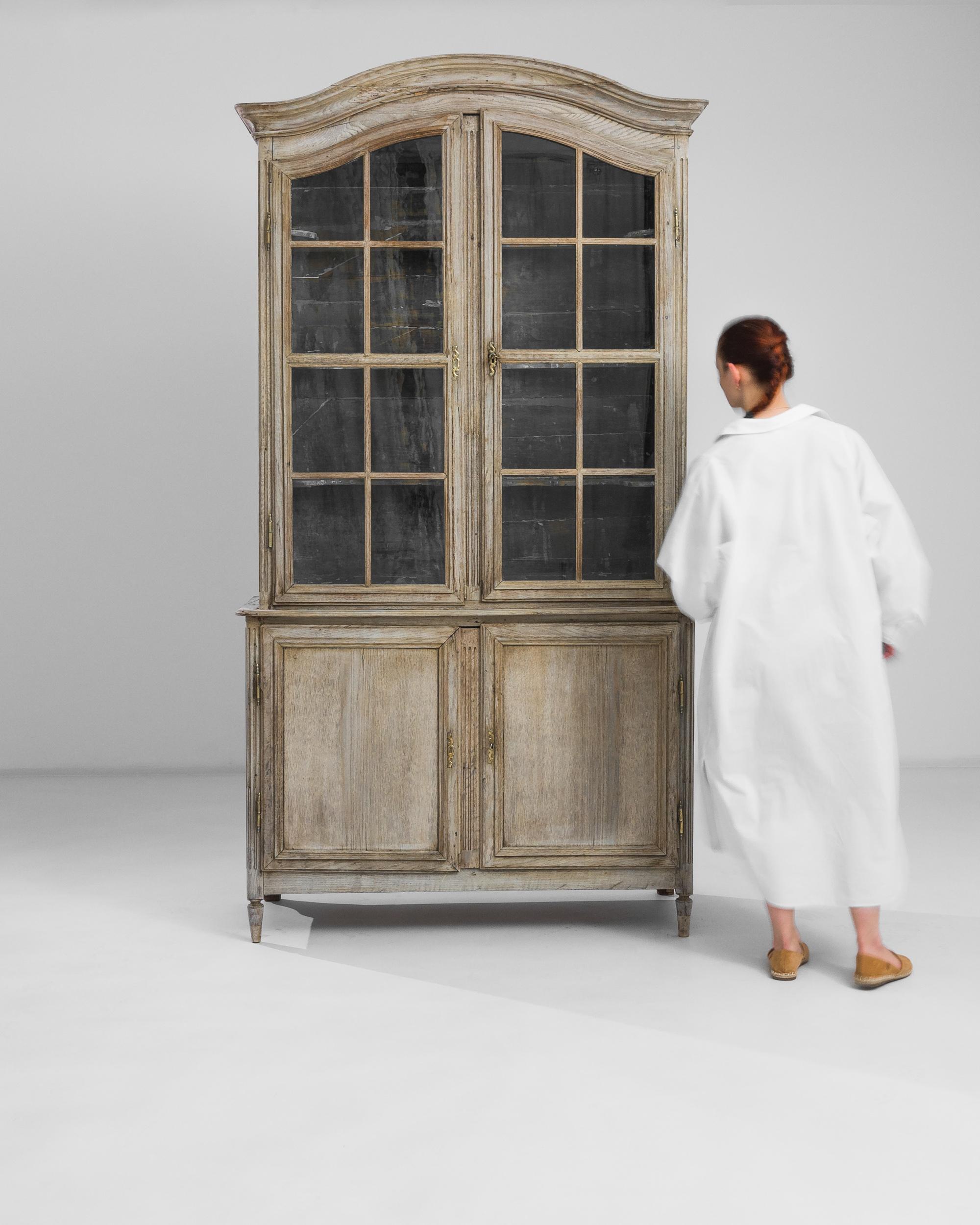This antique oak vitrine offers a display case which is a treasure in its own right. Made in France in the 1860s, the arch of the cabinet cuts an elegant silhouette; mullioned windows give a view onto patinated shelves. The contrast between bright