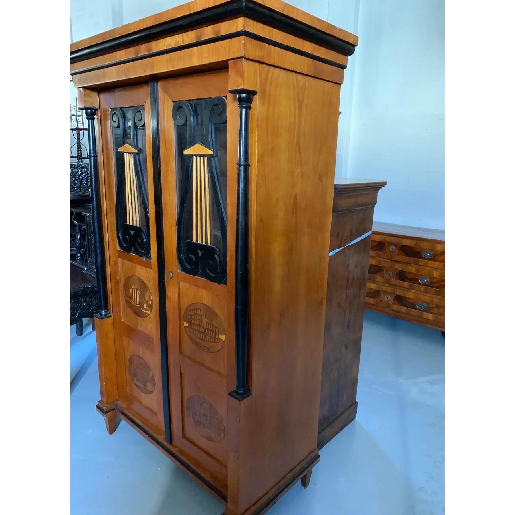 Un armario italiano de estilo Biedermeier de hacia 1860. Dos puertas de armario tienen paneles superiores de cristal cubiertos de liras ebonizadas talladas. Debajo hay paneles empotrados decorados con óvalos incrustados que representan ruinas