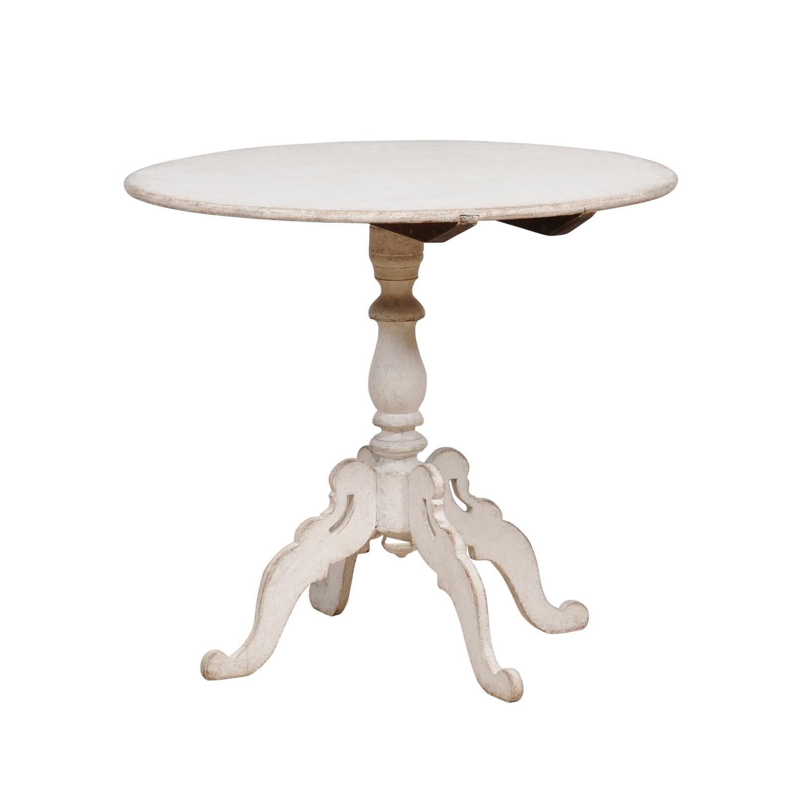 A Swedish light grey painted tilt-top table from circa 1860 with circular top and turned pedestal mounted on four carved scrolling legs. Heralding from circa 1860, this Swedish tilt-top table is a striking blend of functionality and artistry, bathed