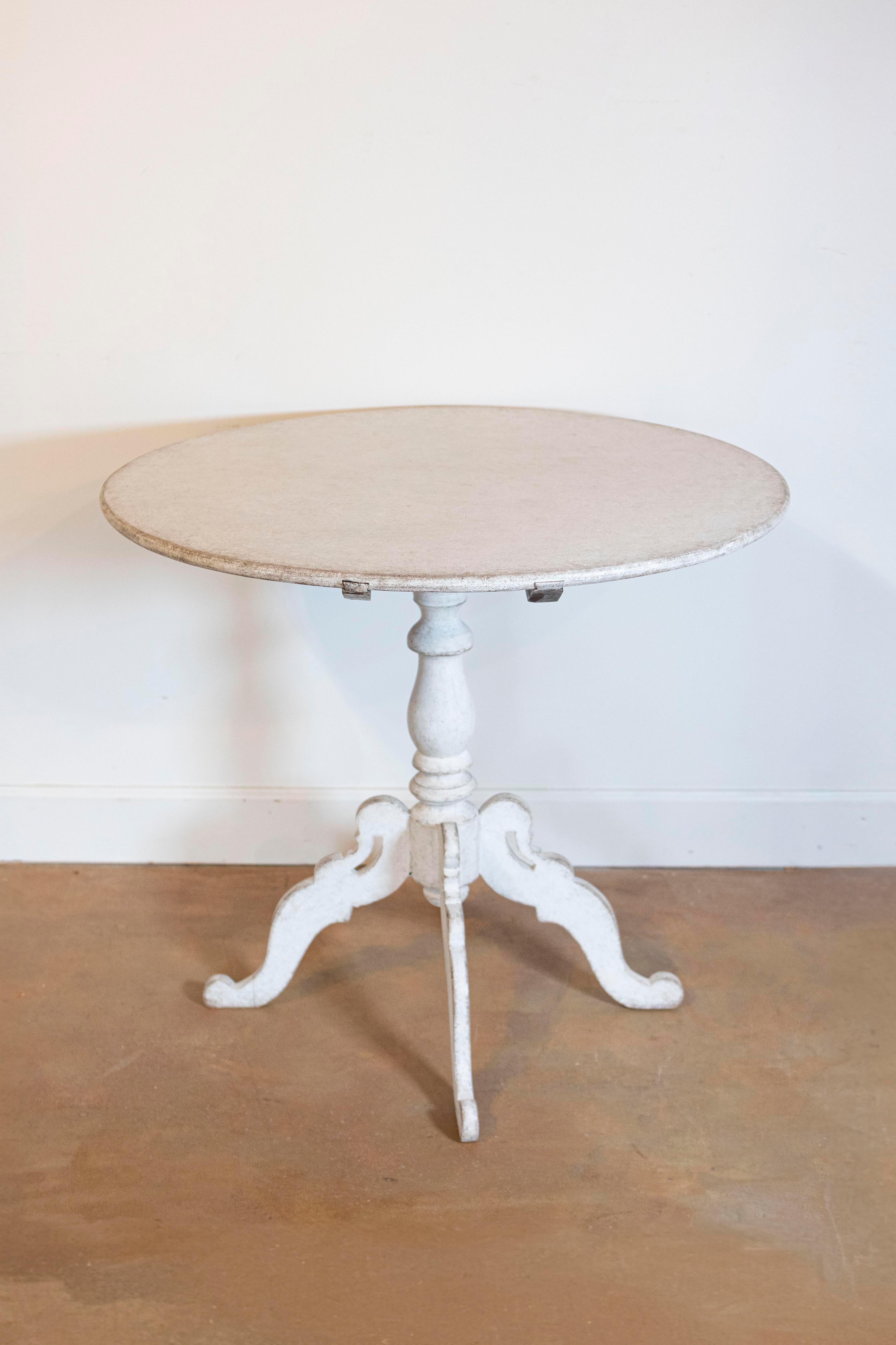 A Swedish light grey painted tilt-top table from circa 1860 with circular top and turned pedestal mounted on four carved scrolling legs. Heralding from circa 1860, this Swedish tilt-top table is a striking blend of functionality and artistry, bathed