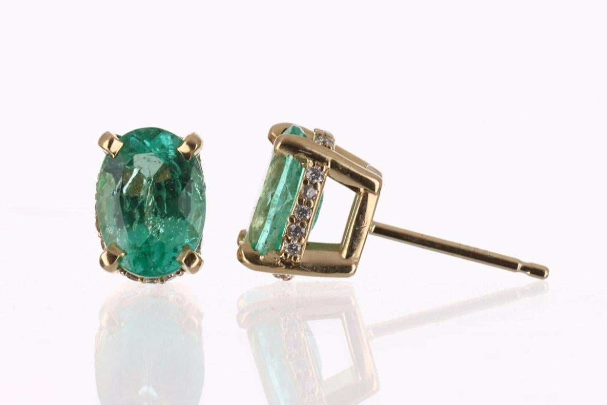 Featured here is a stunning set of oval emerald studs in fine 14K yellow gold with diamond accents along the sides. Displayed are medium-green emeralds with good transparency, accented by diamonds in gold earring mount, allowing for the emerald to