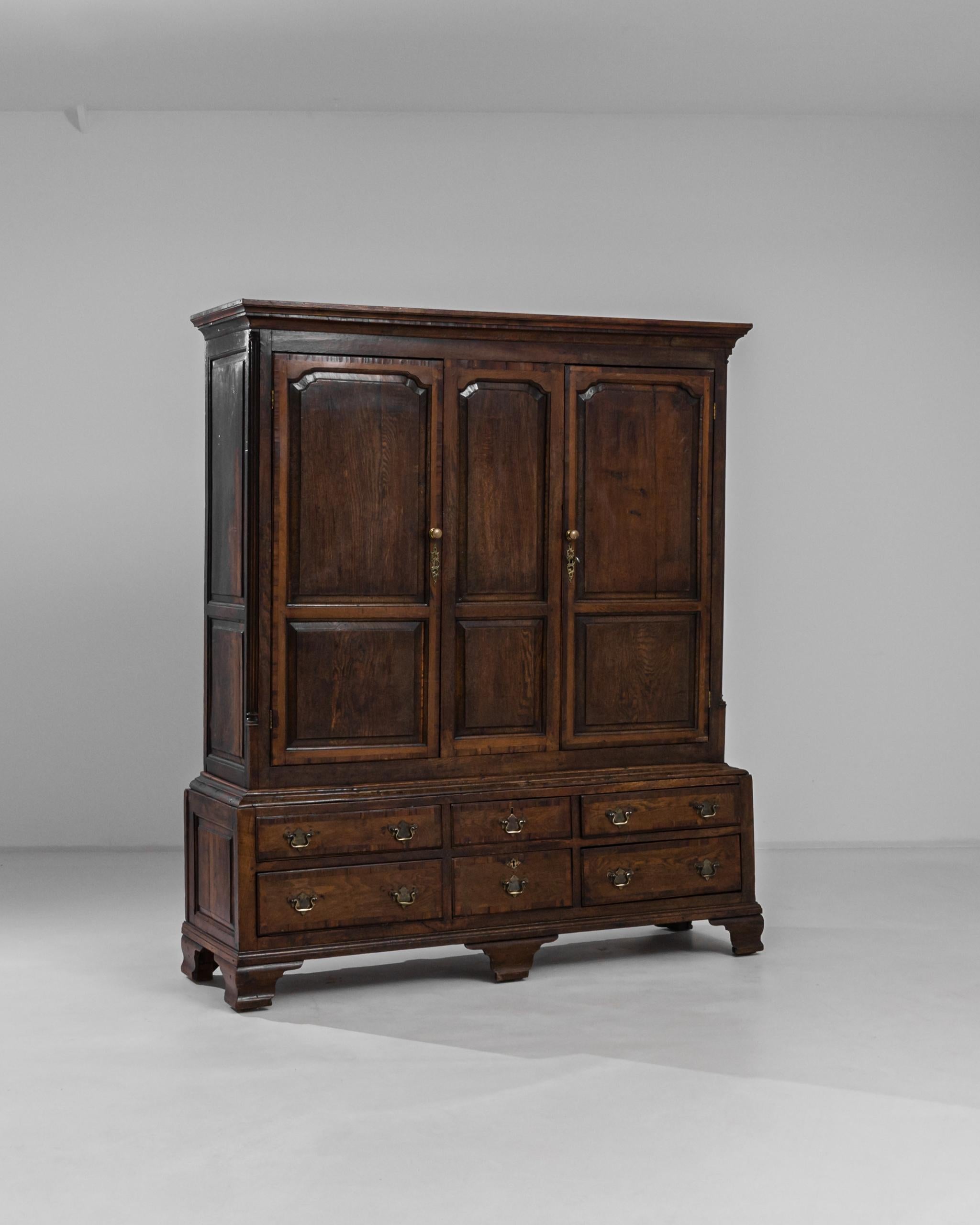 A dark wooden cabinet with brass hardware, circa 1870s England. Dark and warm oak panels comprise a large and elaborate structure. Lined with numerous drawers faced with an arch motif, this cabinet offers a sumptuous storage piece that will elevate