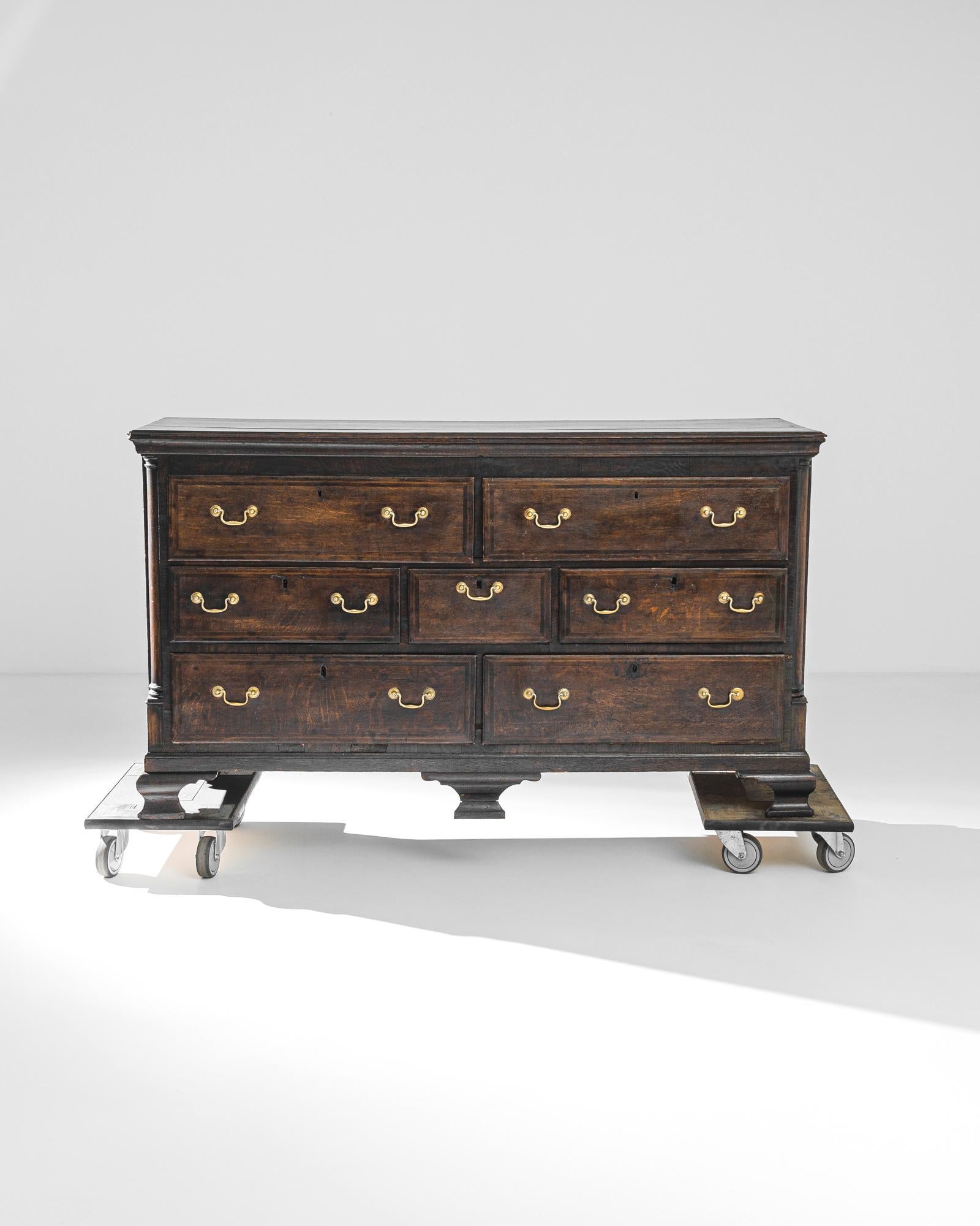 A wooden chest of drawers from the United Kingdom, produced circa 1870. This stately chest of drawers, from late Victorian England, features seven sliding drawers with hanging brass pulls that contrast gorgeously against the refined dark brown wood