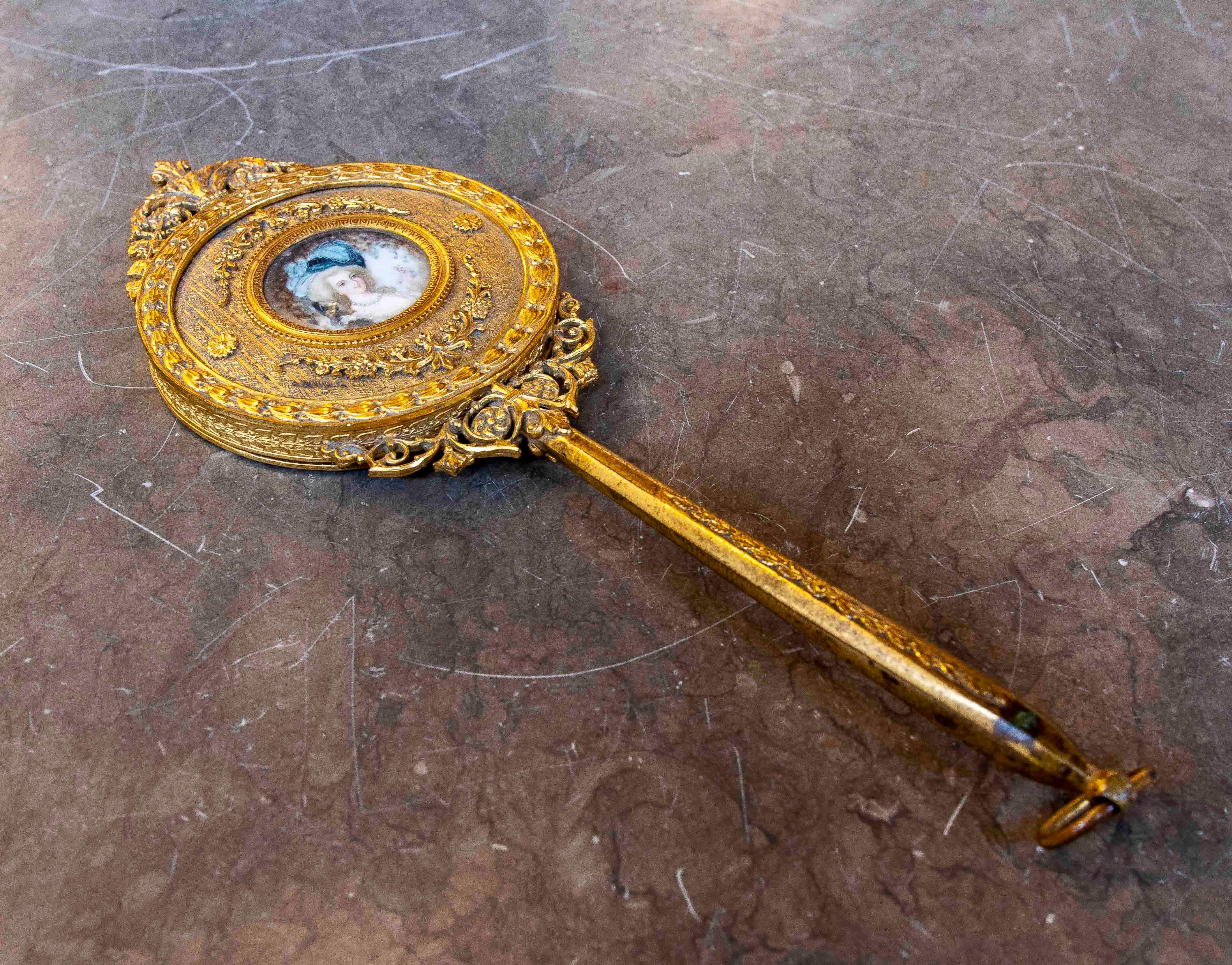 This exquisite hand mirror features a gold frame richly decorated with floral motifs and ornamental details. Includes a painted miniature in the center of the porcelain reverse, depicting a precisely framed portrait. The piece reflects a baroque
