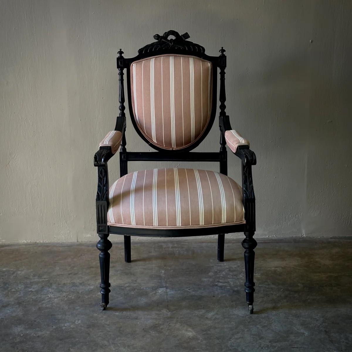 This Louis XVI style fauteuil open armchair radiates opulence with its masterfully carved ebonized wood and the custom taupe and cream striped linen upholstery. The neoclassical carvings, reminiscent of laurel wreaths and acanthus leaves, enhance