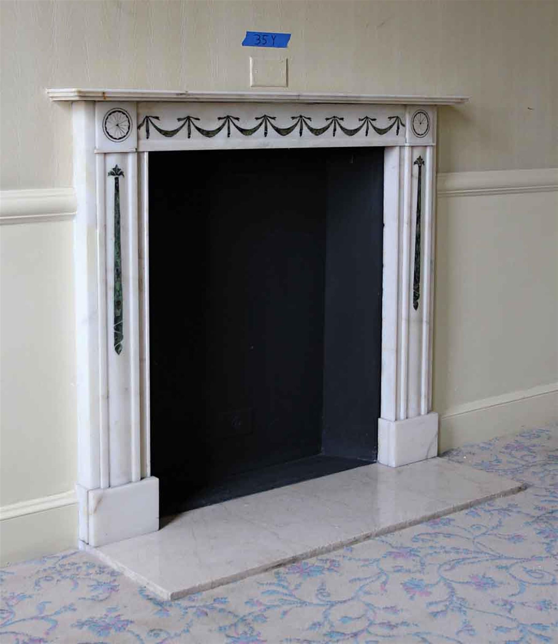 A small scale white marble mantel captures attention with its exquisite detailing. The mantel boasts an exceptional green inlay that has been remarkably well-preserved over time. This inlay is a true work of art, intricate and finely crafted. While