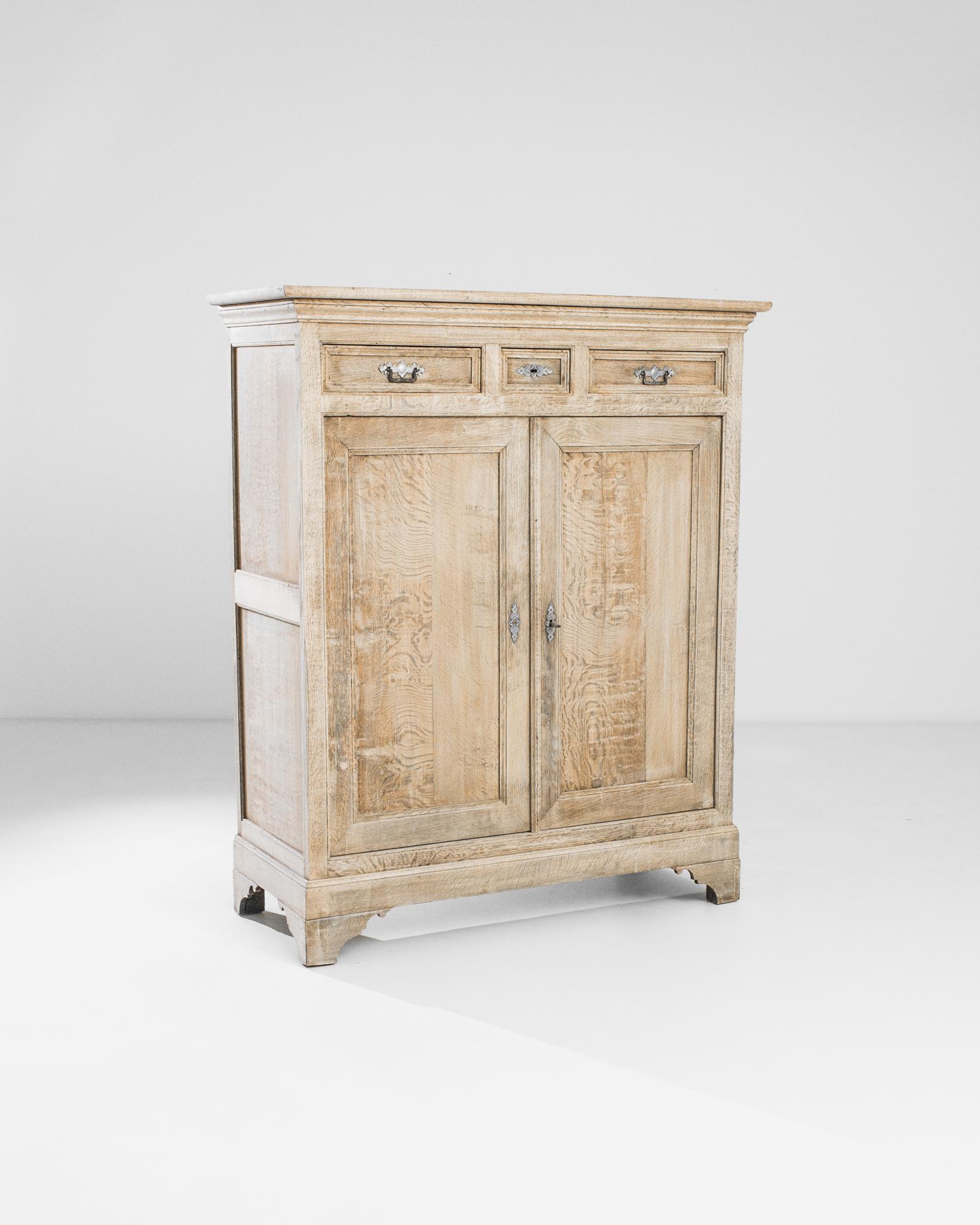 An oak cabinet from 1880s Belgium. Scalloped feet lend the upright case a poised stance. The restoration of the oak showcases its natural beauty — a warm honeyed tone, with a liquid grain. The intricate design of the pewter lock pieces and drawer