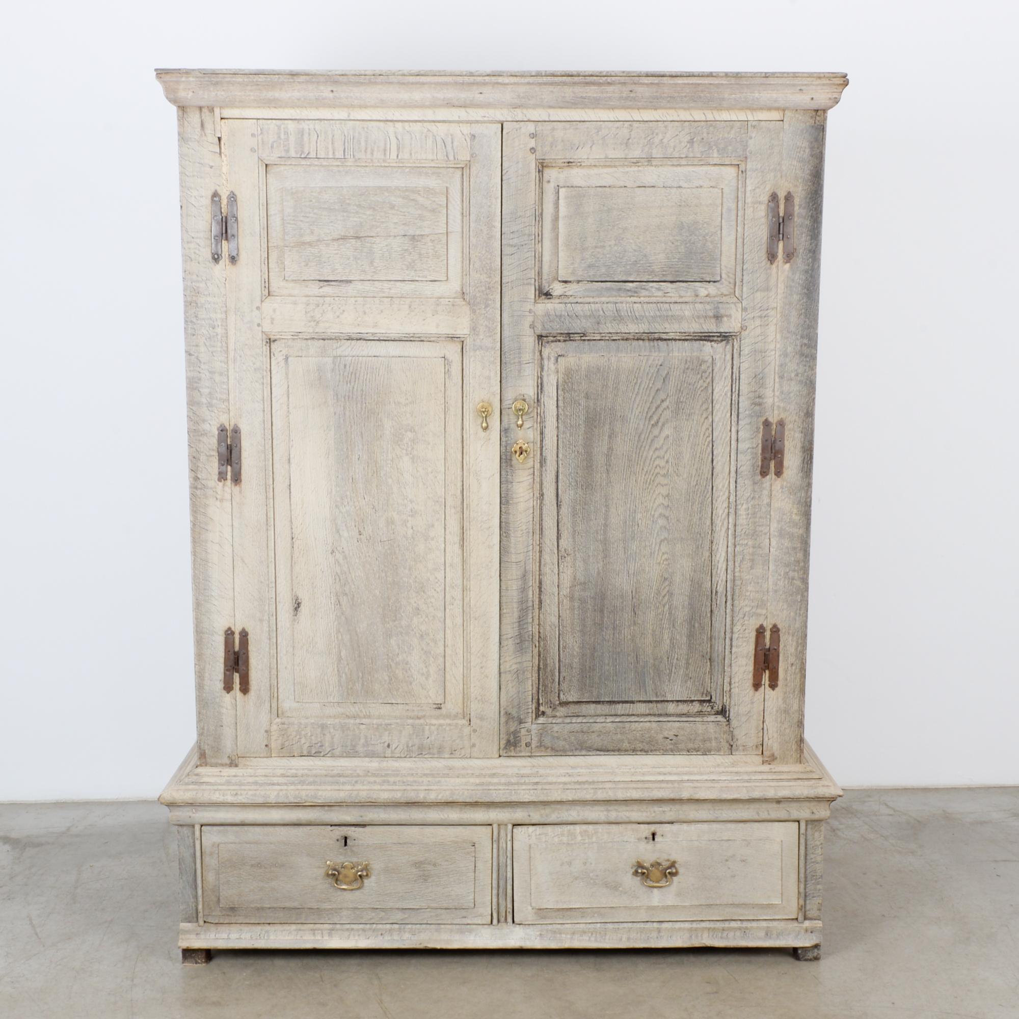 A bleached oak armoire from France, circa 1880. Two paneled doors open onto an interior shelf. Below, two drawers pull out with gilded handles. The square cabinet is enhanced by original details: butterfly hinges create an attractive border; gilded