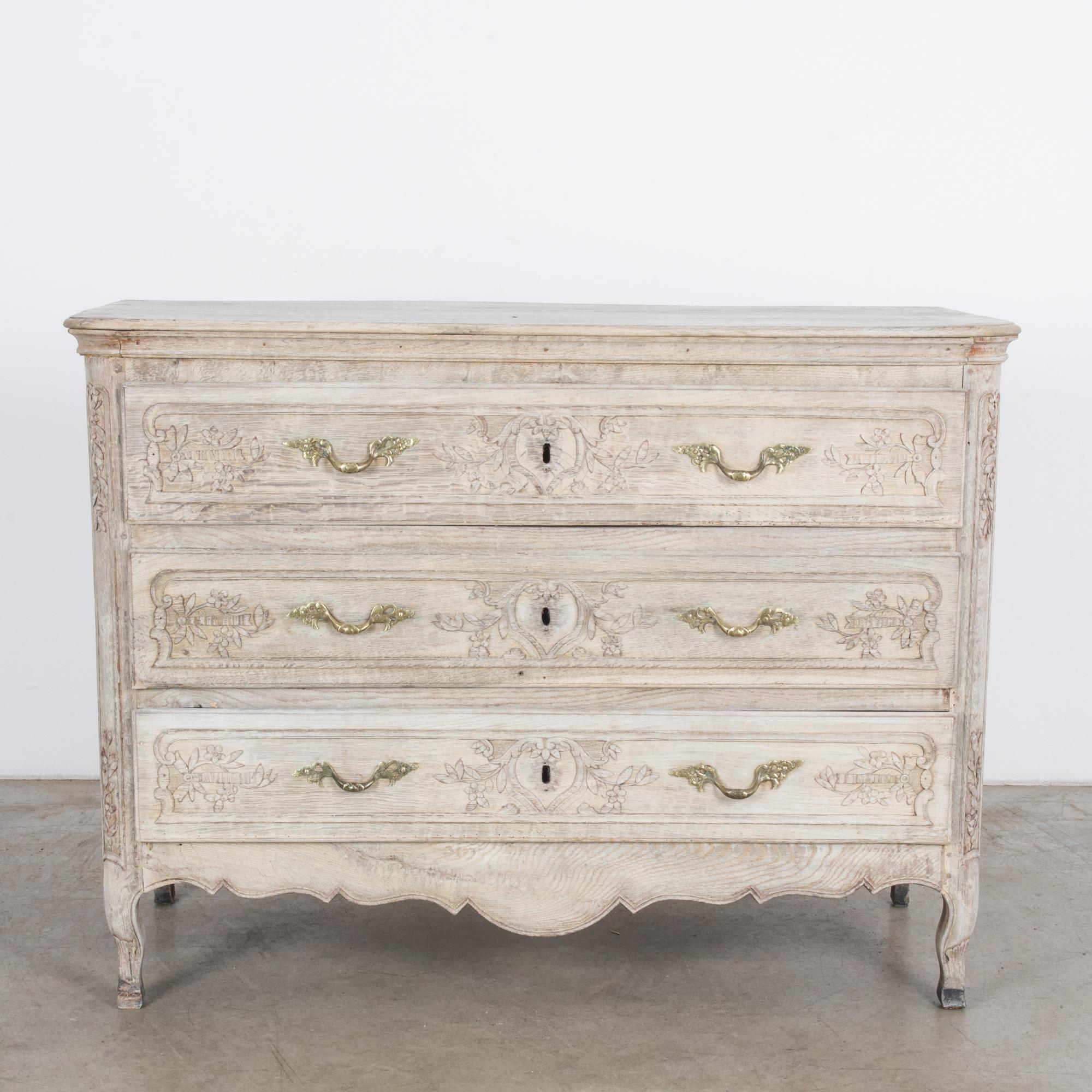 Made in 1880s France, this captivating bleached oak chest of drawers displays cabriole legs and carvings, popular features of the French Provincial style. Intricate floral and foliage carvings on the three drawers and front corners lend an old-world