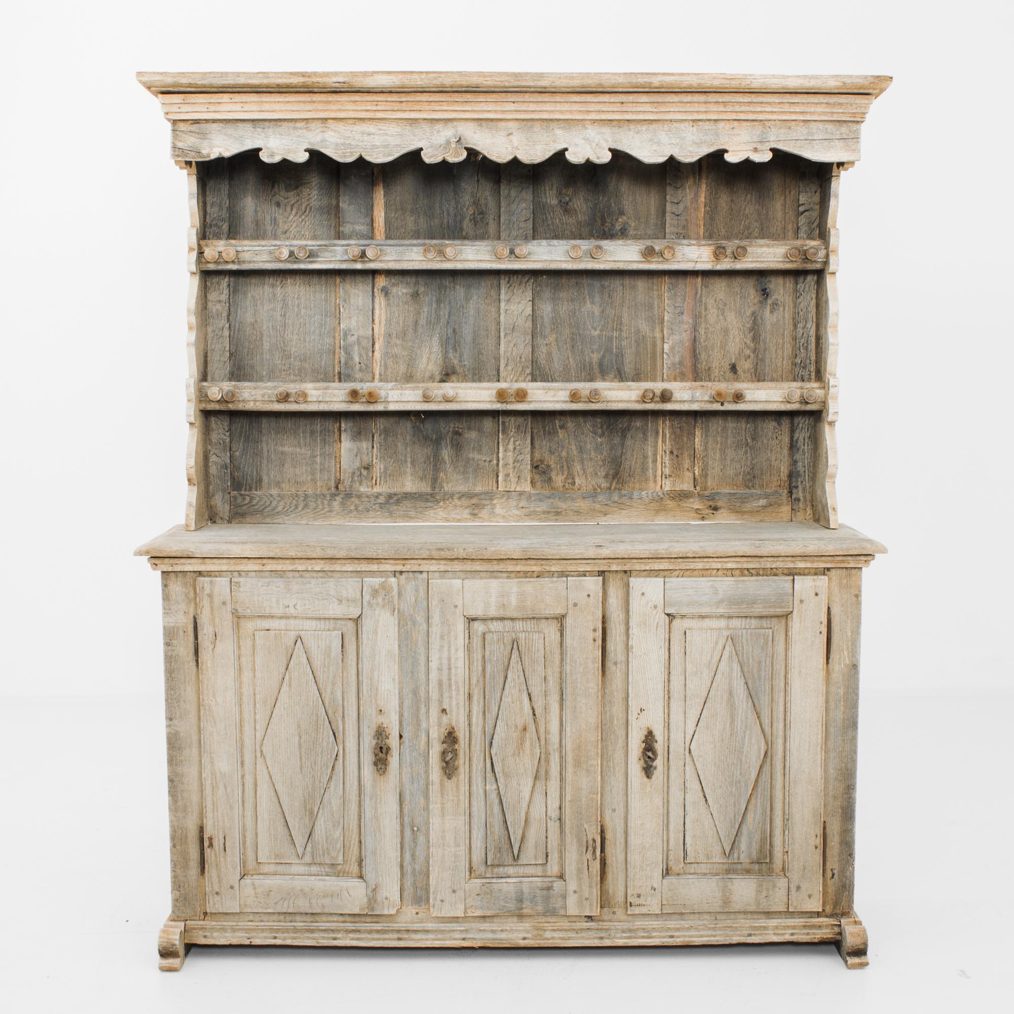 An oak dresser from 1880s France with a charming country aesthetic. A carved canopy frames the upper section, equipped with rails of round wooden knobs for hanging hang mugs, ornaments or bundles of herbs. Carved diamonds on the panelling of the