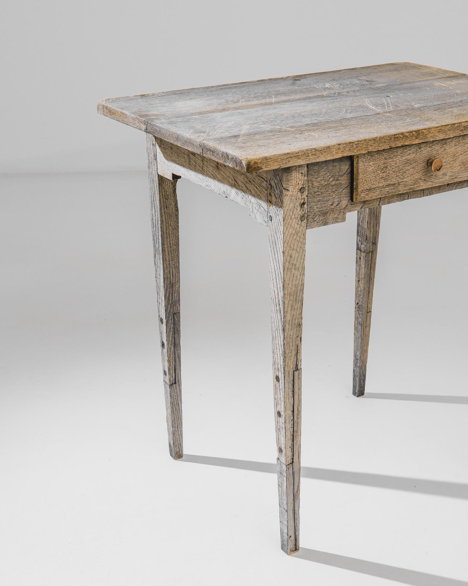 A bleached oak table from France produced circa 1880. Featuring a single sliding drawer tucked into a deep apron underneath a gray plain of a tabletop, this leggy little occasional table is ready for action. Each leg is extended with a stilt