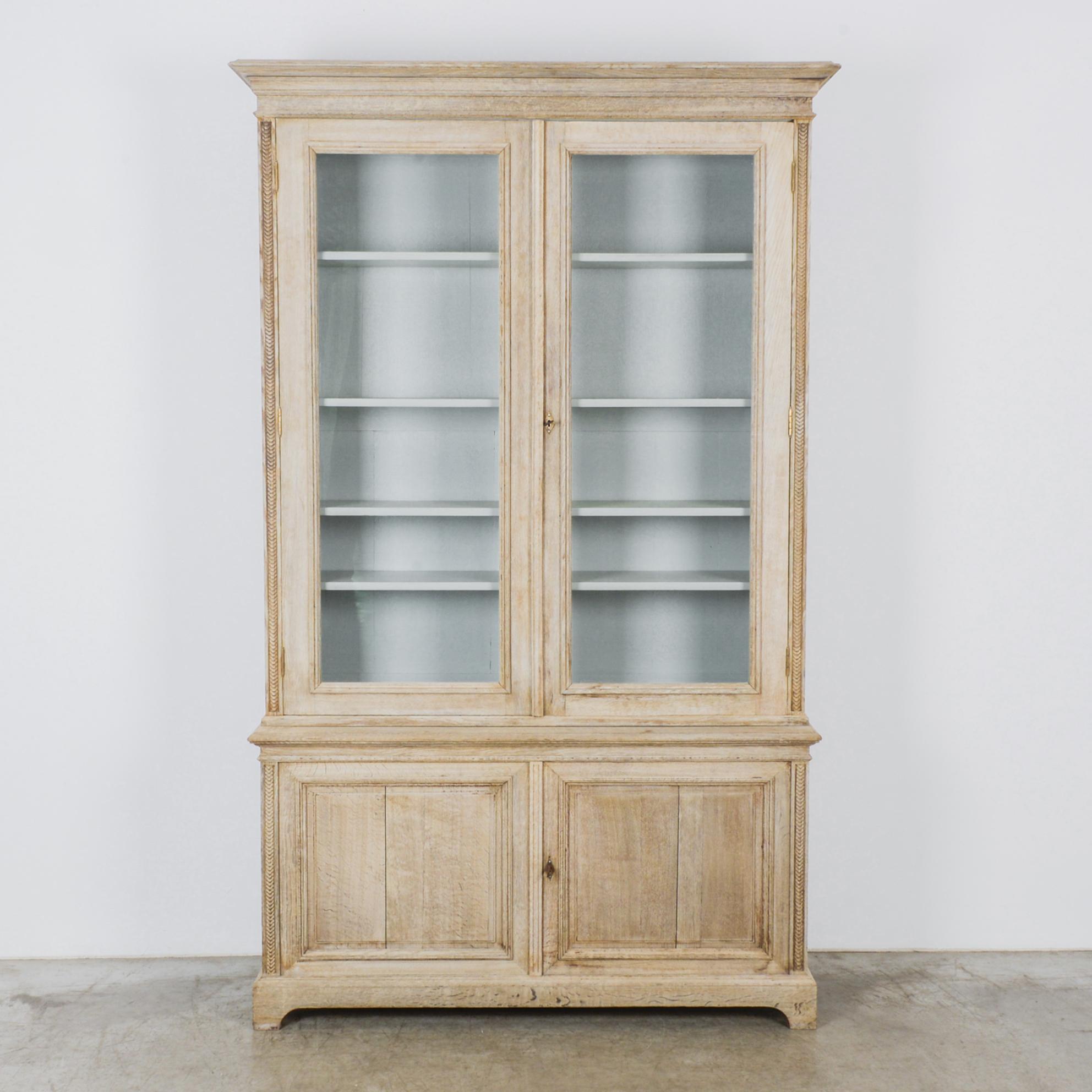 A bleached oak vitrine from France, circa 1880s. Glass fronted double doors open onto four shelves. The interior is painted a robin’s egg blue, complementing the light, cheerful tone of the bleached oak. Beneath the vitrine is a small cabinet with a
