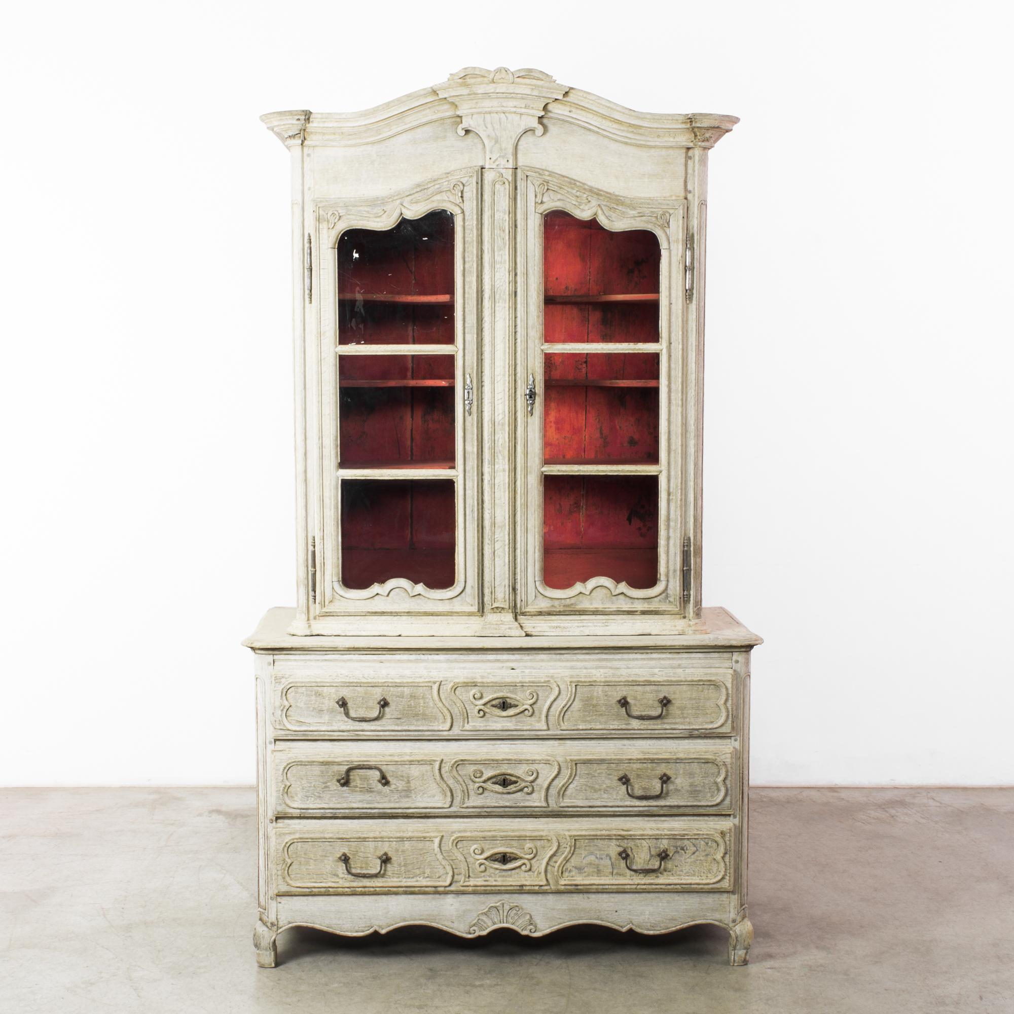 Made in 1880s France, this two-part bleached oak vitrine features a delightful crimson interior, setting off the exquisite wood grain and smooth finish. The contoured paneling, carved curlicues, and eye-like carvings on the drawers evoke the rich