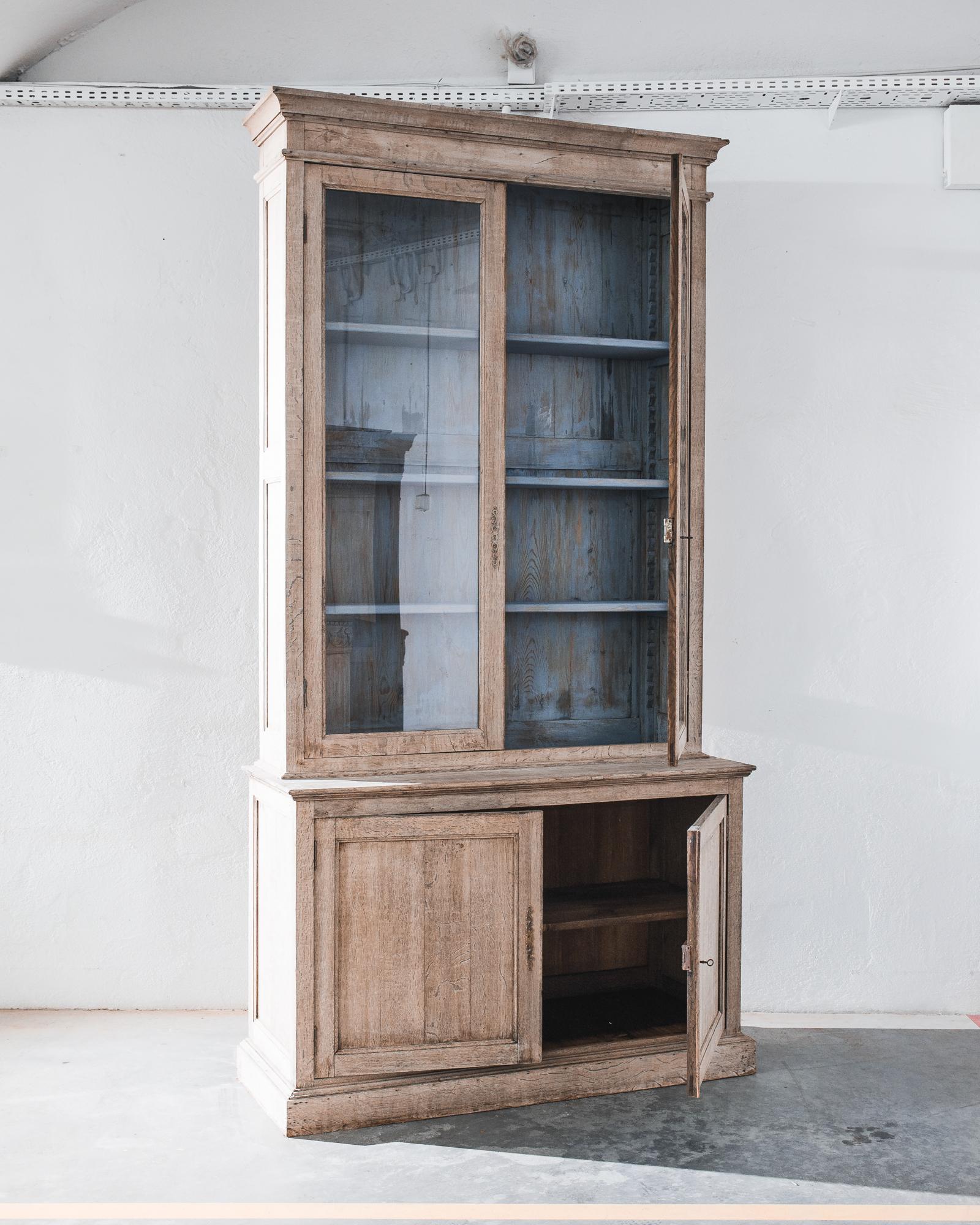 A bleached oak vitrine from France, produced circa 1880. A grand vitrine standing nine feet tall, featuring two levels of cabinets: an upper story fronted by glass with a gray patinated interior of four shelves, and a lower double cabinet of two
