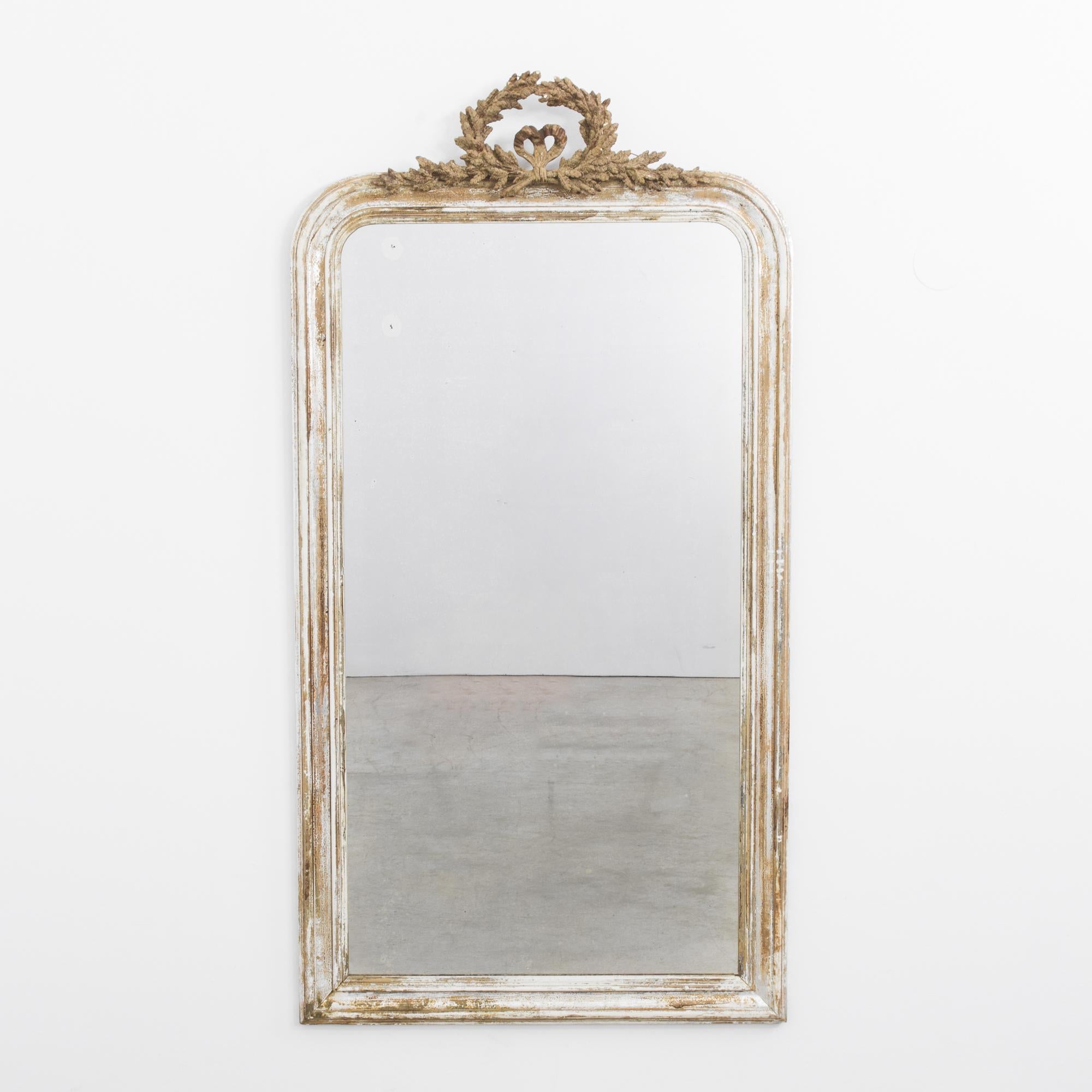 A gilded wooden mirror from France, circa 1880, with a romantic patina of fresh white and muted gold. Tall and stately, the curve of the upper corners of the frame creates an arched silhouette, crowned by a carved laurel wreath. Elegant and