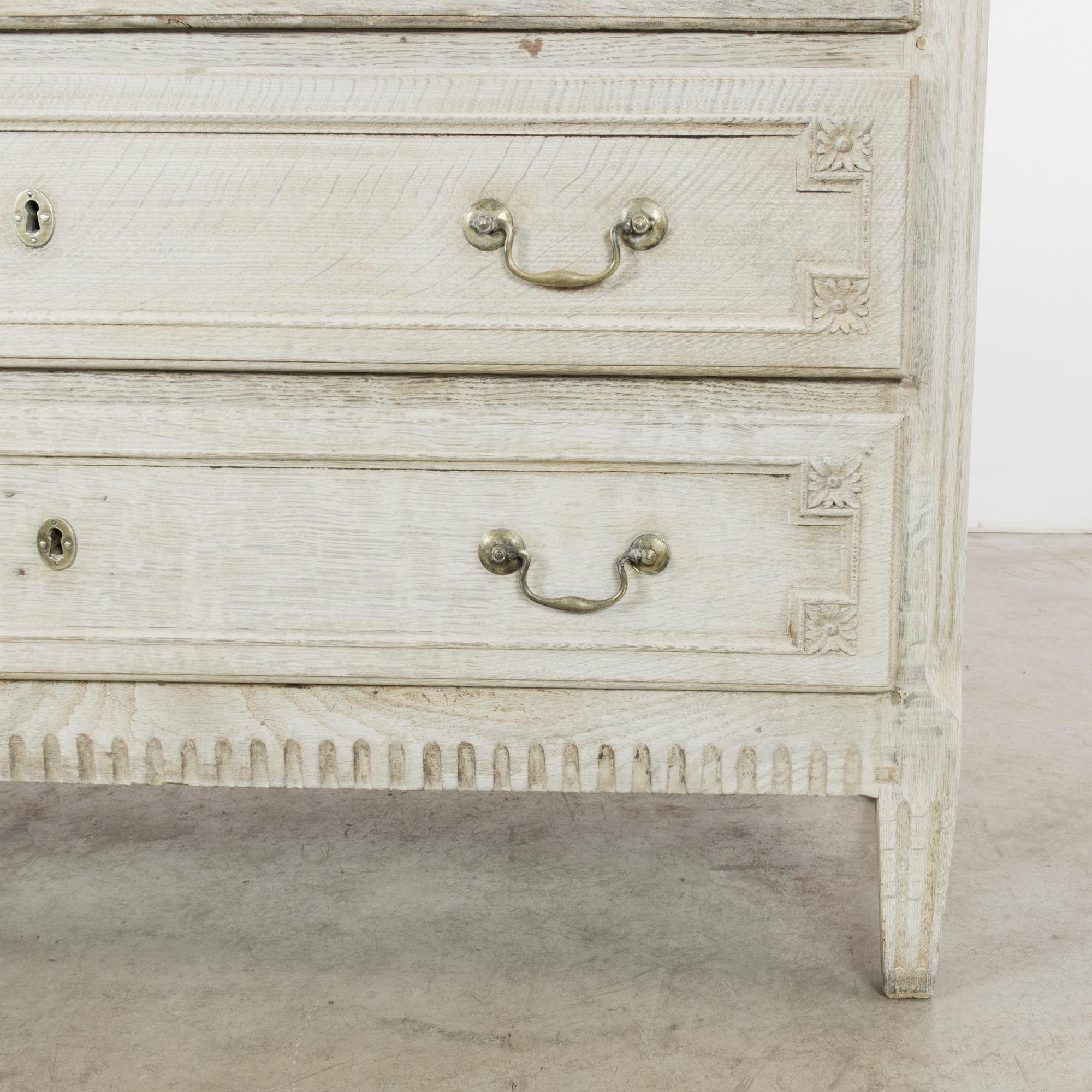 An antique chest of drawers from 1880s France. The oak has been restored to an ivory finish; gilded drop handles bring out the delicate tone. The upright shape is enhanced by elegant Neoclassical ornament. Carved rosettes frame the panels of the