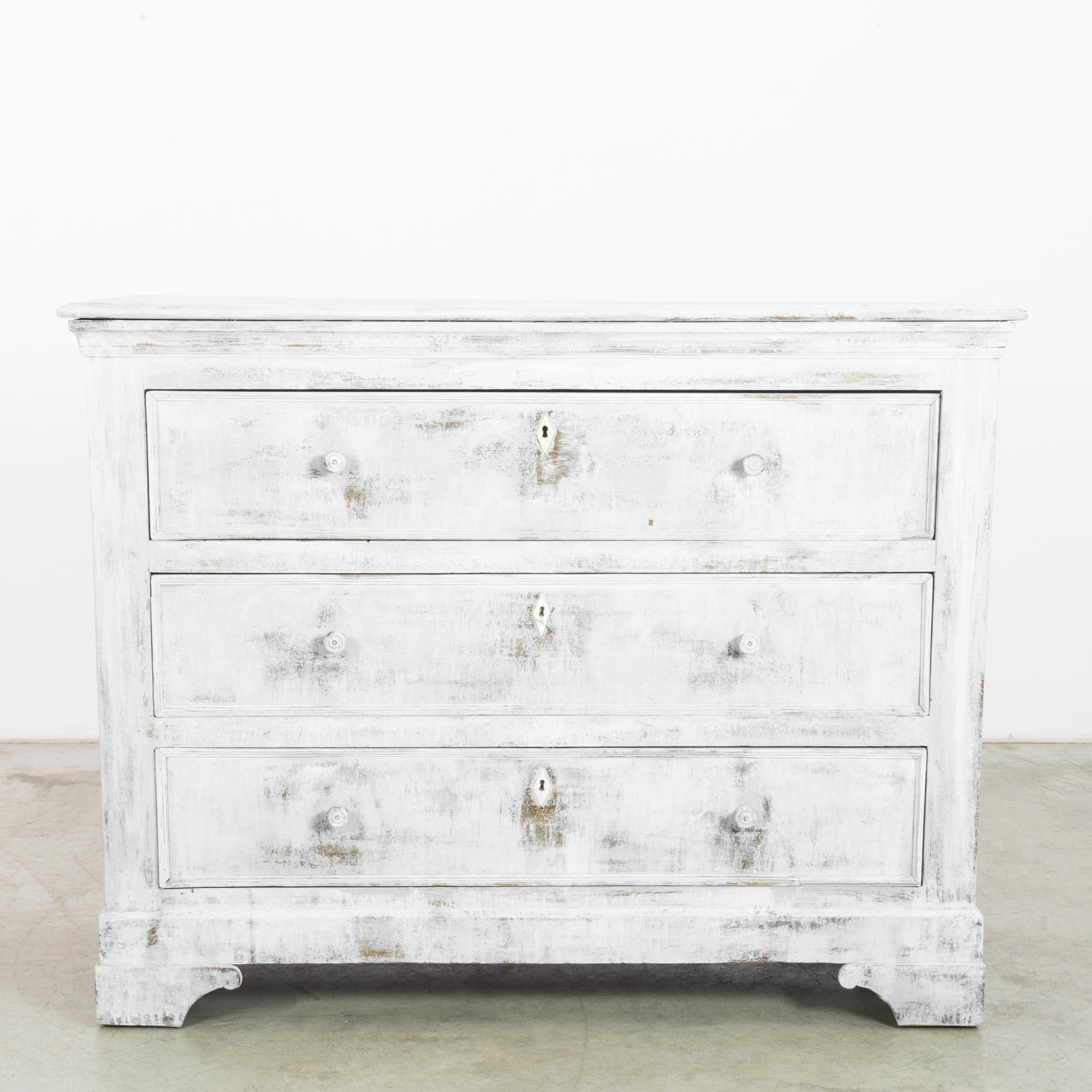 Made in France, circa 1880, this wooden chest of drawers displays a simple geometric silhouette, complemented by the crown molding and circular pulls. Bracket feet provide a slight elevation. With its distressed milky white patina, it evokes the