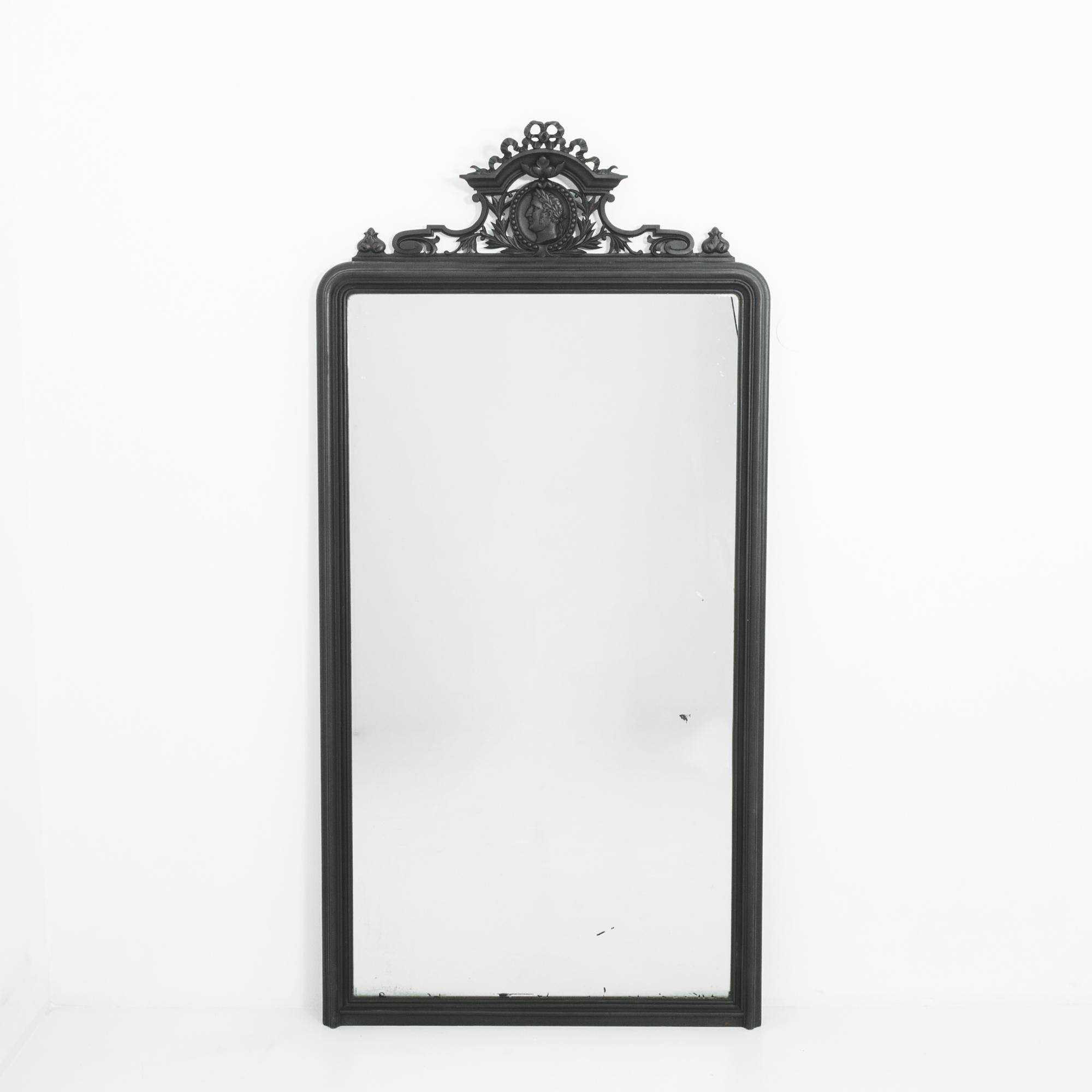 A wooden framed mirror, produced in France in the 1880s. The stark outline of the black frame lends to the commanding presence of this six foot eight inch neoclassical antique. These restrained lines are juxtaposed against an ornate crest of bowed