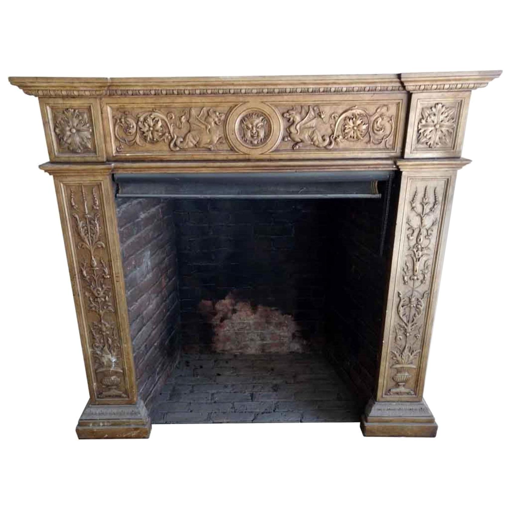 This late 1800s Gothic style fireplace mantel is made of carved gray Carrara marble that has been painted tan. There are floral details with griffins and a face at the center, egg and dart detailing below the shelf, and urns at the base of the front
