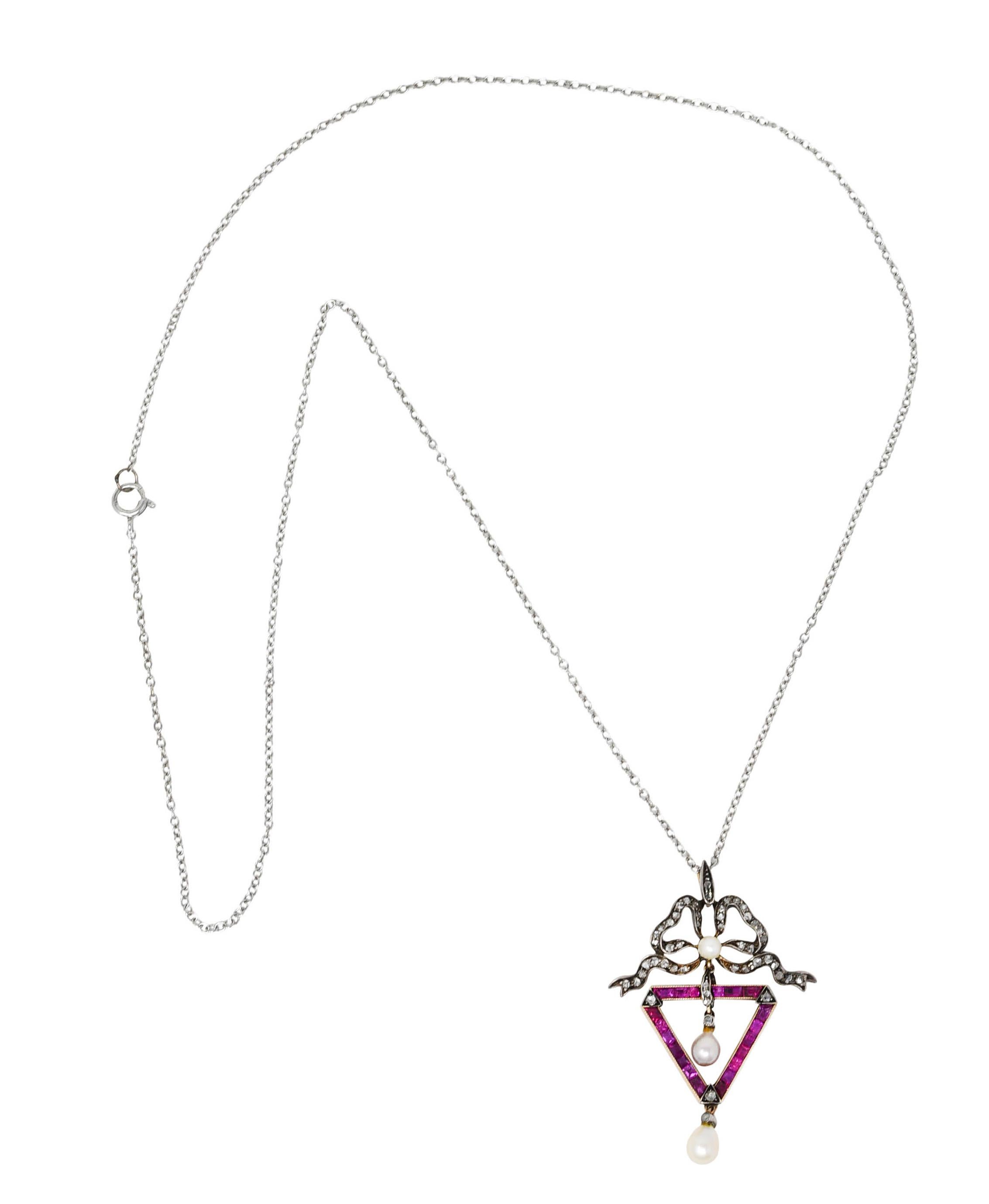 White gold cable chain necklace suspends an articulated pendant with dangling pearl drops. Surmount is a highly dynamic ribboned bow motif that suspends an inverted triangle form. Triangle is channel set with calibrè cut rubies weighing