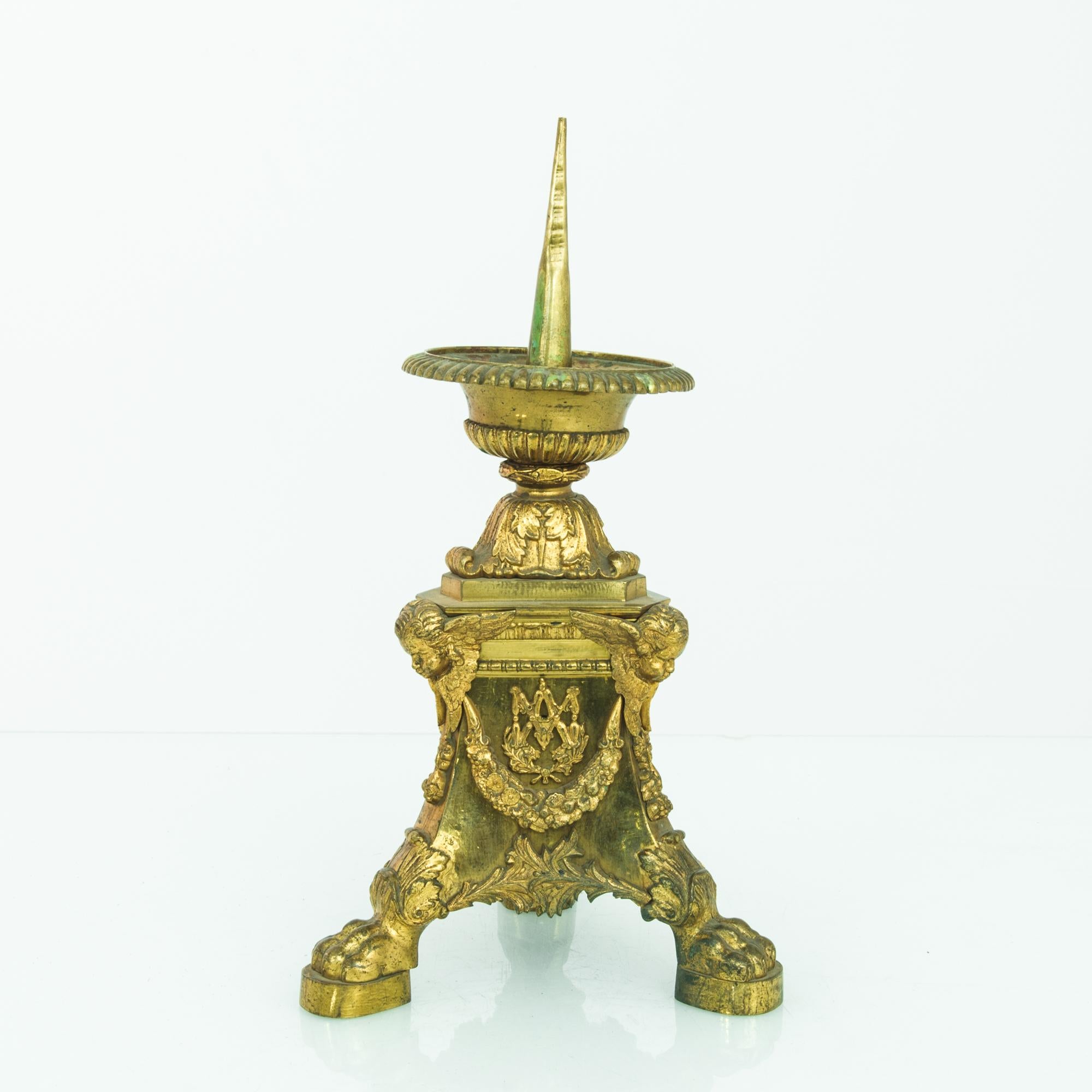 A liturgical candlestick from 1890s France, made of ornate brass. The base is heavily adorned with claw feet, festoons, leafy flourishes and winged cherub heads. Above, a long spike provides a support for a large candle. The bright tone of the brass