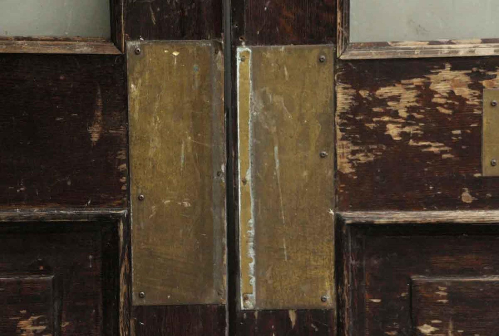 Large 1890s dark wood tone brownstone doors with one vertical center glass panel, two bottom wooden panels and brass kick plates. The opposite side of the doors have one push bar. Priced as a pair. Measures: 100.5 in. H x 60 in. W.