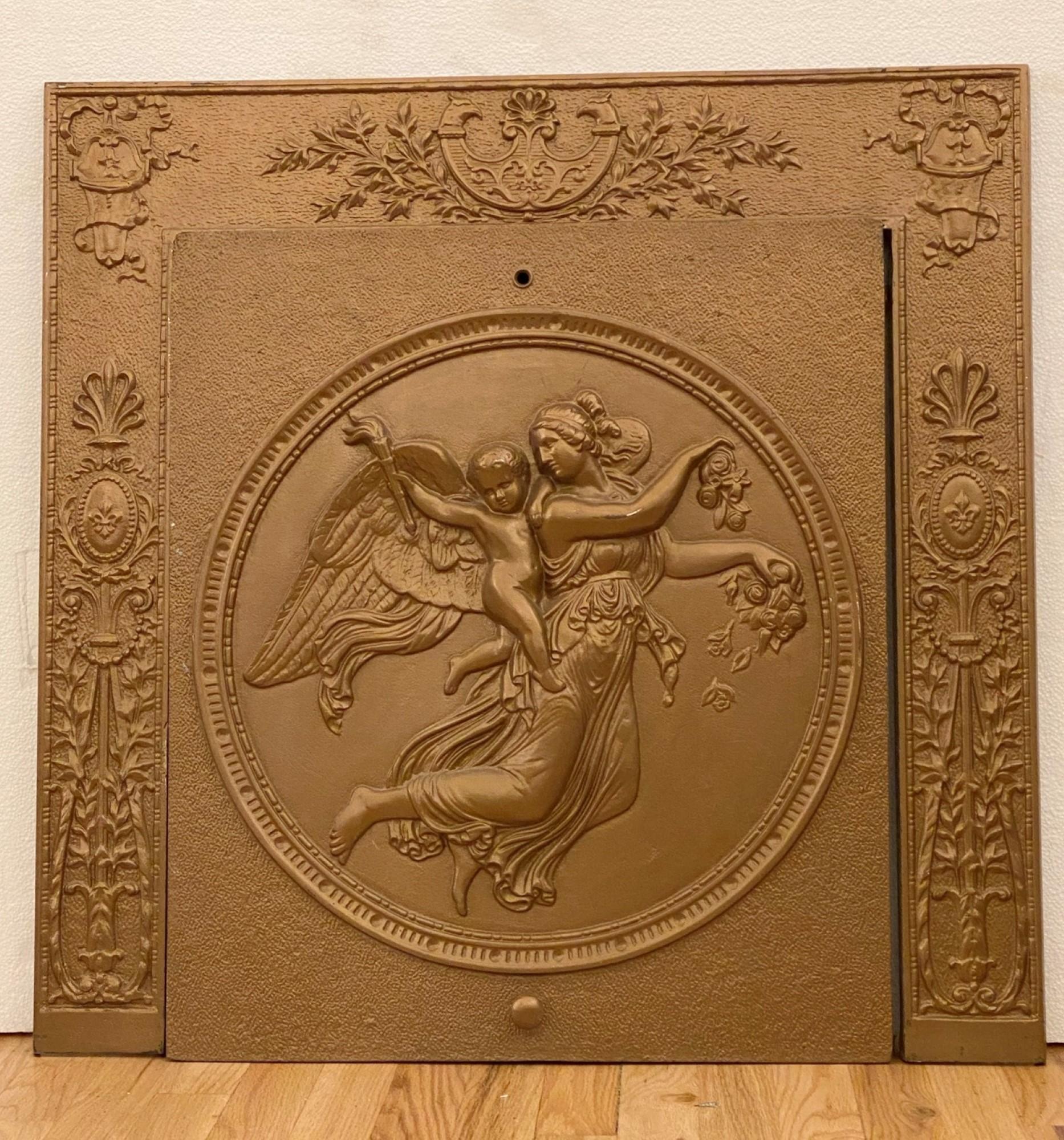 1880s Victorian era summer fireplace cover painted in dark gold with a woman and cherub motif. The Victorian clothed woman is sprinkling flowers with the cherub on her shoulder protects her with a torch in hand. This can be seen at our 333 West