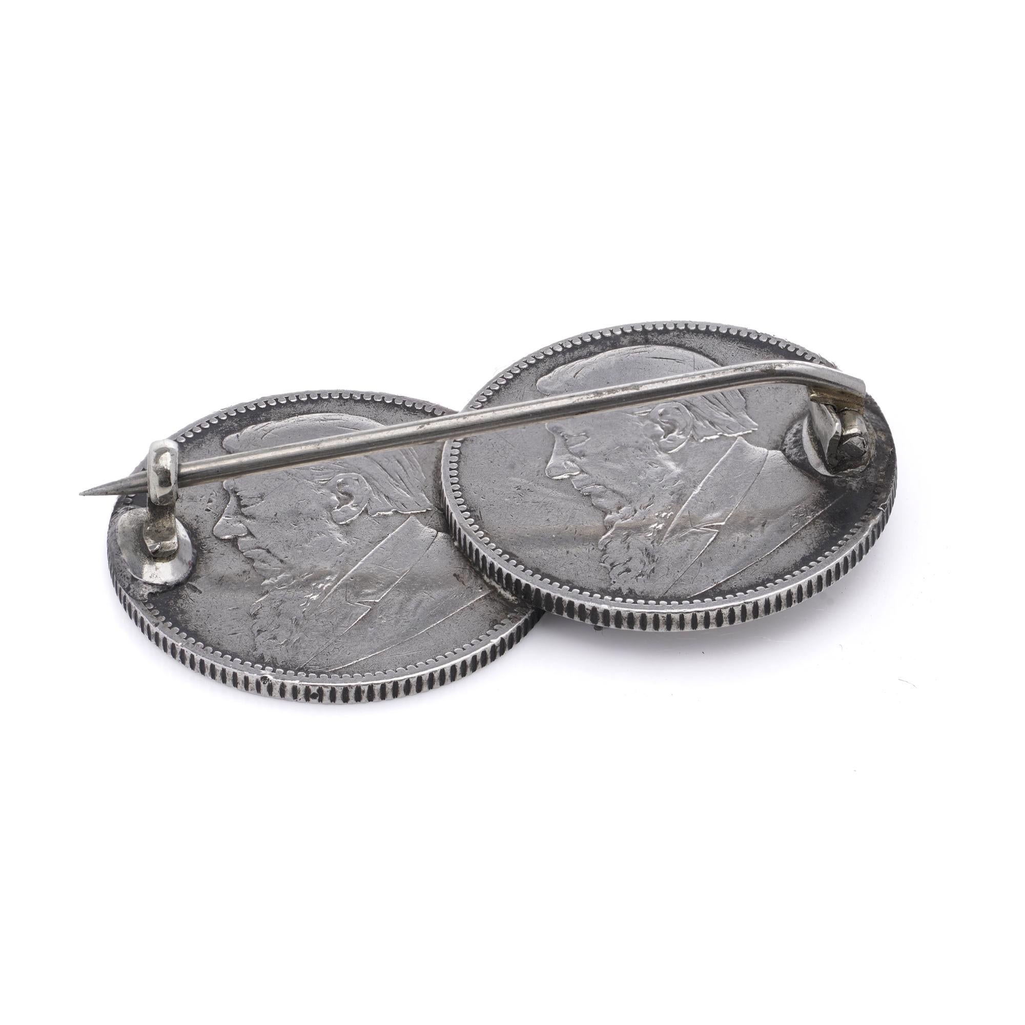 This brooch is made from two 1896 South African Republic (ZAR) 1 Shilling coins, featuring the profile of Paul Kruger, president of the ZAR. The coins are made of silver (0.925 fine) and were minted during a period of economic prosperity fueled by