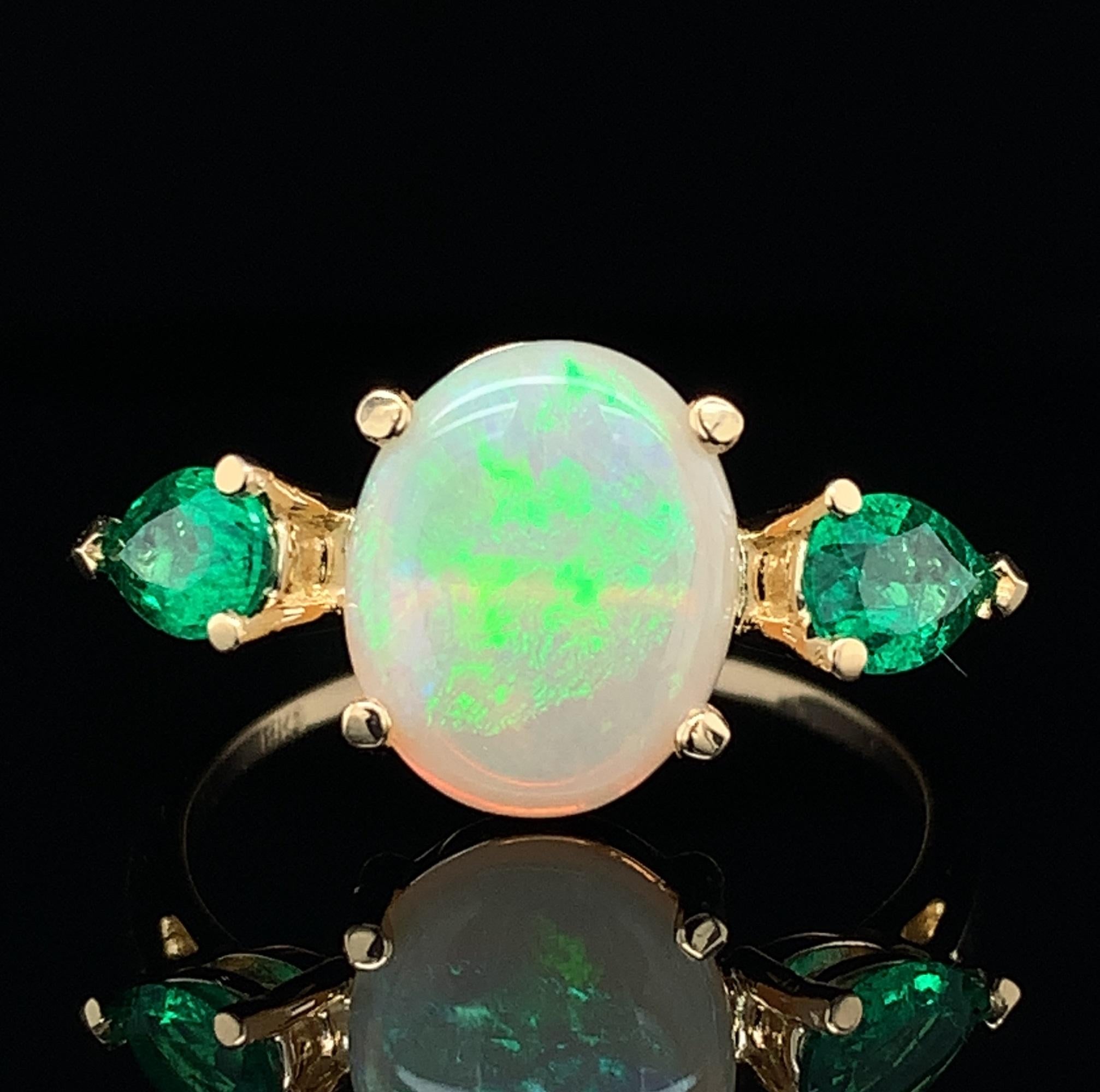 18K yellow gold ring featuring a 1.76 carat  opal with emeralds. The opal has lively play of color on a white background with green and yellow.  The oval opal measures about 11mm x 9mm. There are 2 pear shaped genuine earth mined emeralds weighing