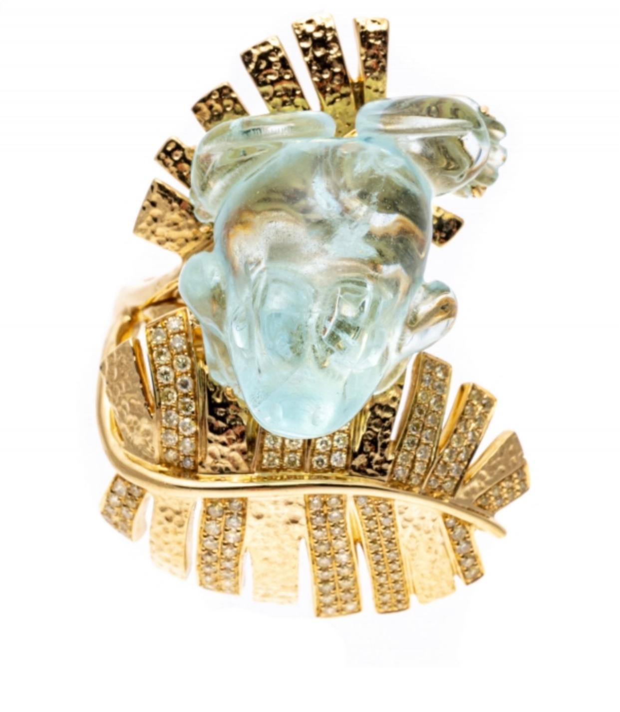 This magnificent ring features a carved, pale greenish blue color aquamarine carved in the shape of a frog; sitting atop fringed tropical leaf fronds of patterned yellow gold and alternating fronds set with yellow and white round faceted diamonds,