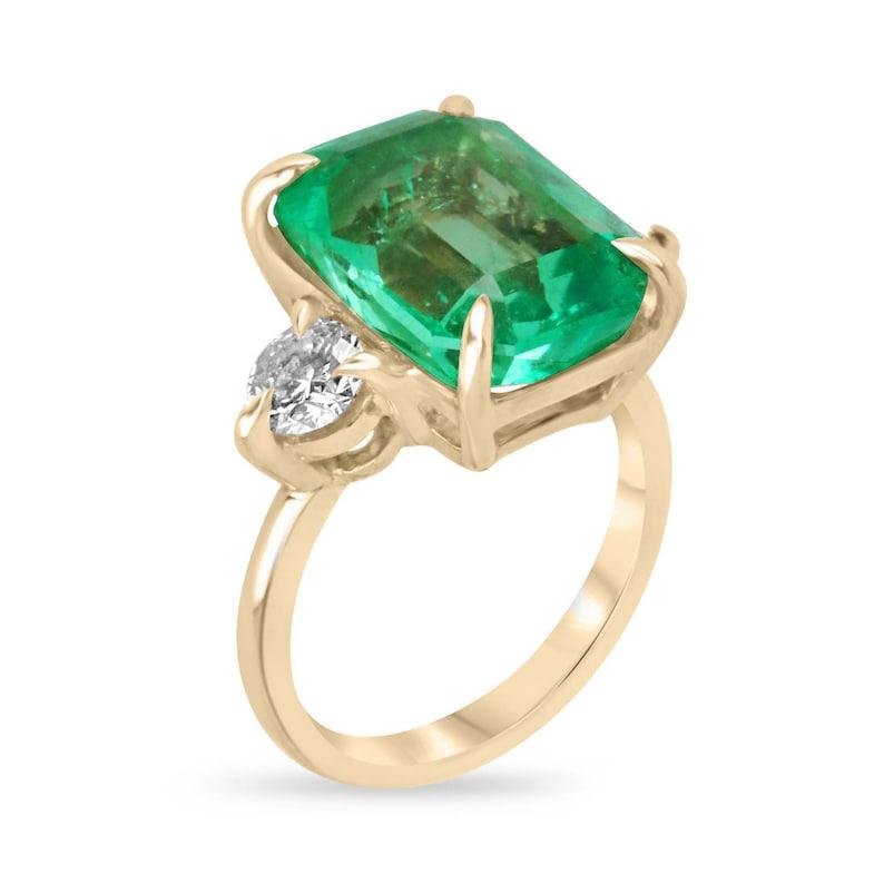 Behold the elegance of our Emerald and Diamond Three Stone Ring, featuring a stunning emerald-cut emerald set north to south in a refined four-claw prong setting. The emerald boasts a vivid medium dark green hue, with strong saturation and tone,