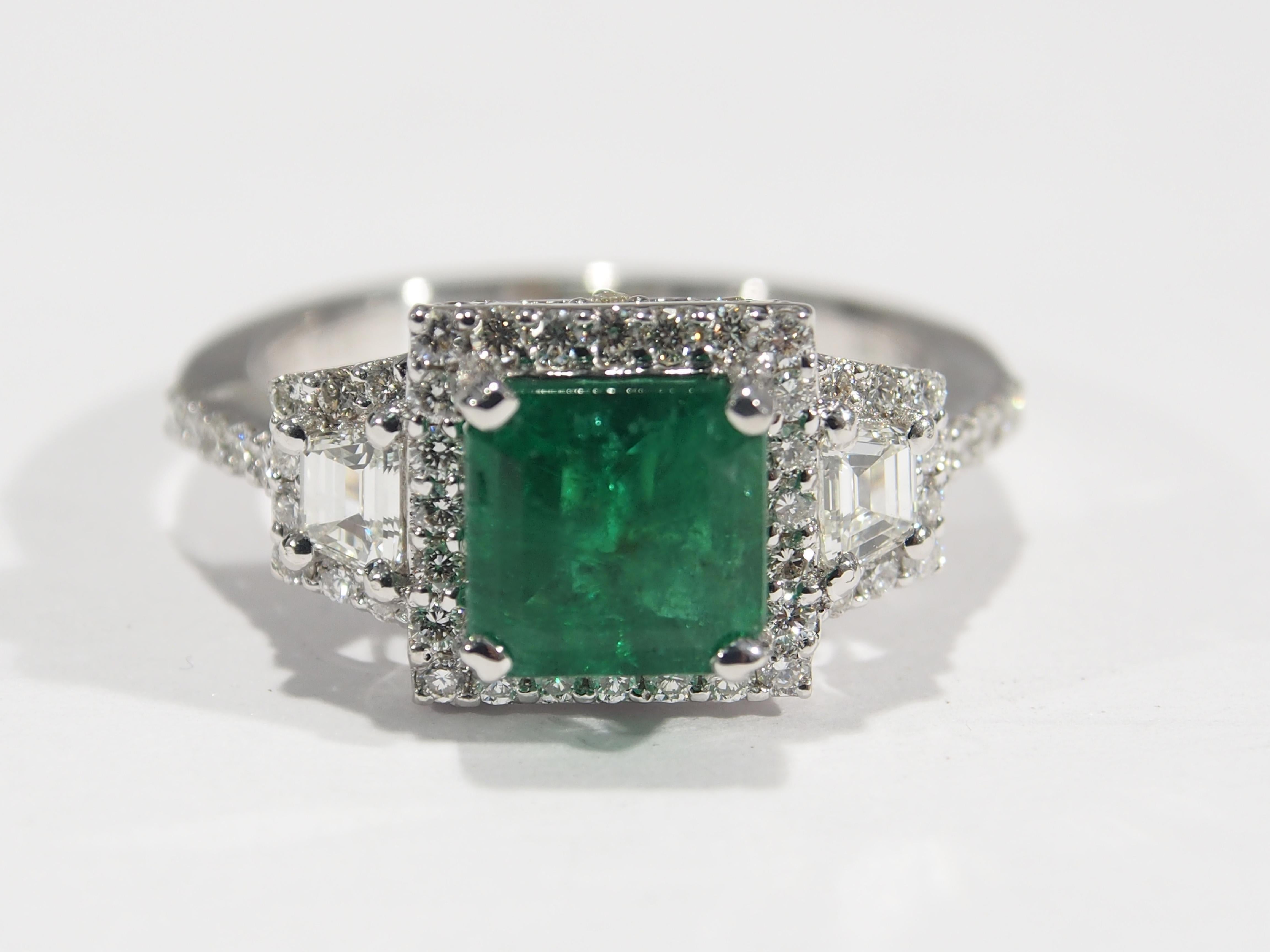A beautiful Natalie K. 18K white gold three stone ring featuring a 1.5ct Emerald with (2) Trapezoid Diamonds and (92) Round Brilliant Cut diamonds surround the Emerald and side Trapezoids. The diamonds are approximately .82 total weight, G-I in