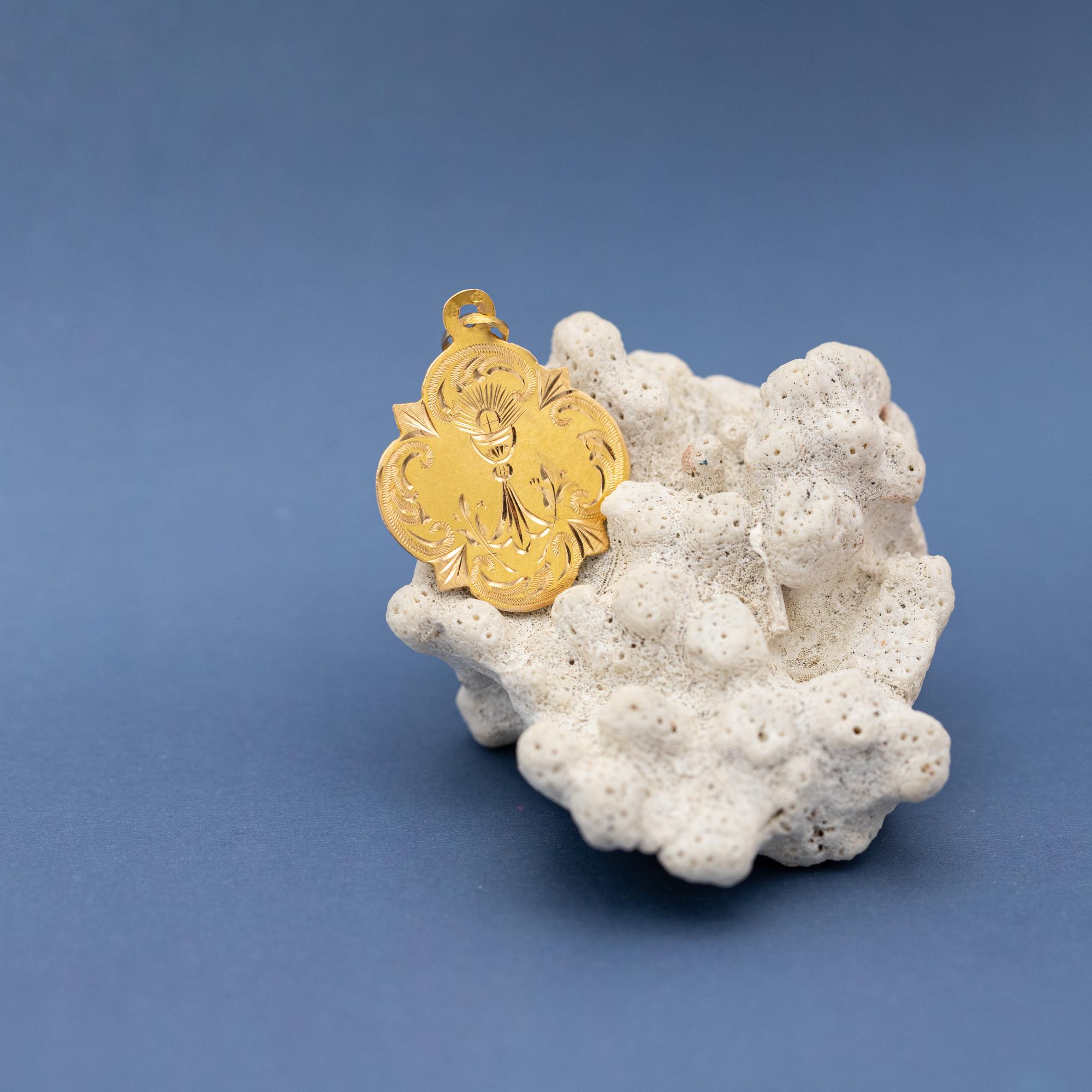 Nous avons le plaisir de vous présenter cette jolie médaille patinée en or jaune 18 ct représentant un calice, coupe souvent utilisée pour boire lors d'une cérémonie religieuse. Une hostie marquée d'une croix scintille au-dessus d'elle. Ce pendentif