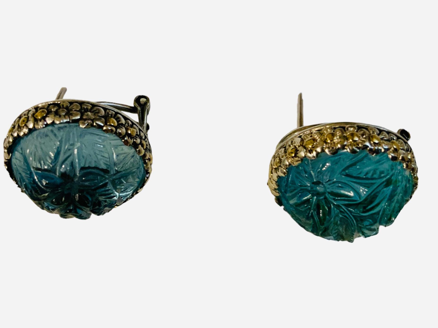 This is an 18K Gold and 925 Sterling Silver Blue Topaz pair of earrings. It depicts a pair of earrings consisting of a light blue topaz round dome carved with branches of flowers and leaves mounted in 925 sterling bezel setting. The border around