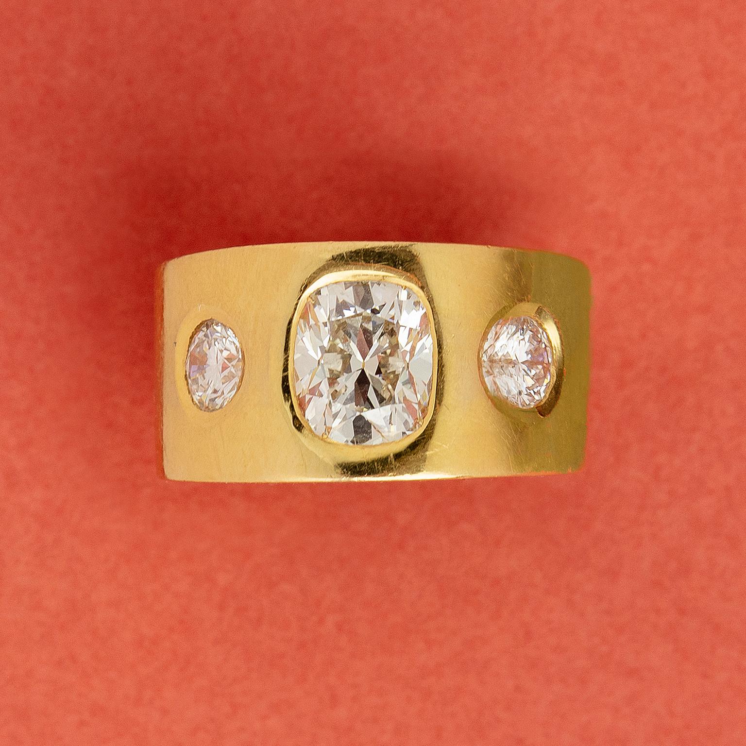 An 18-carat gold, bulky, three-stone band ring with sharp edges set with a large cushion-cut diamond (app. 2.6 carats, J, VS2) and two brilliant-cut diamonds (each app. 0.50 carat, F-G, VS2-SI1) all in a flush setting. 
weight: 19.46 grams
size: