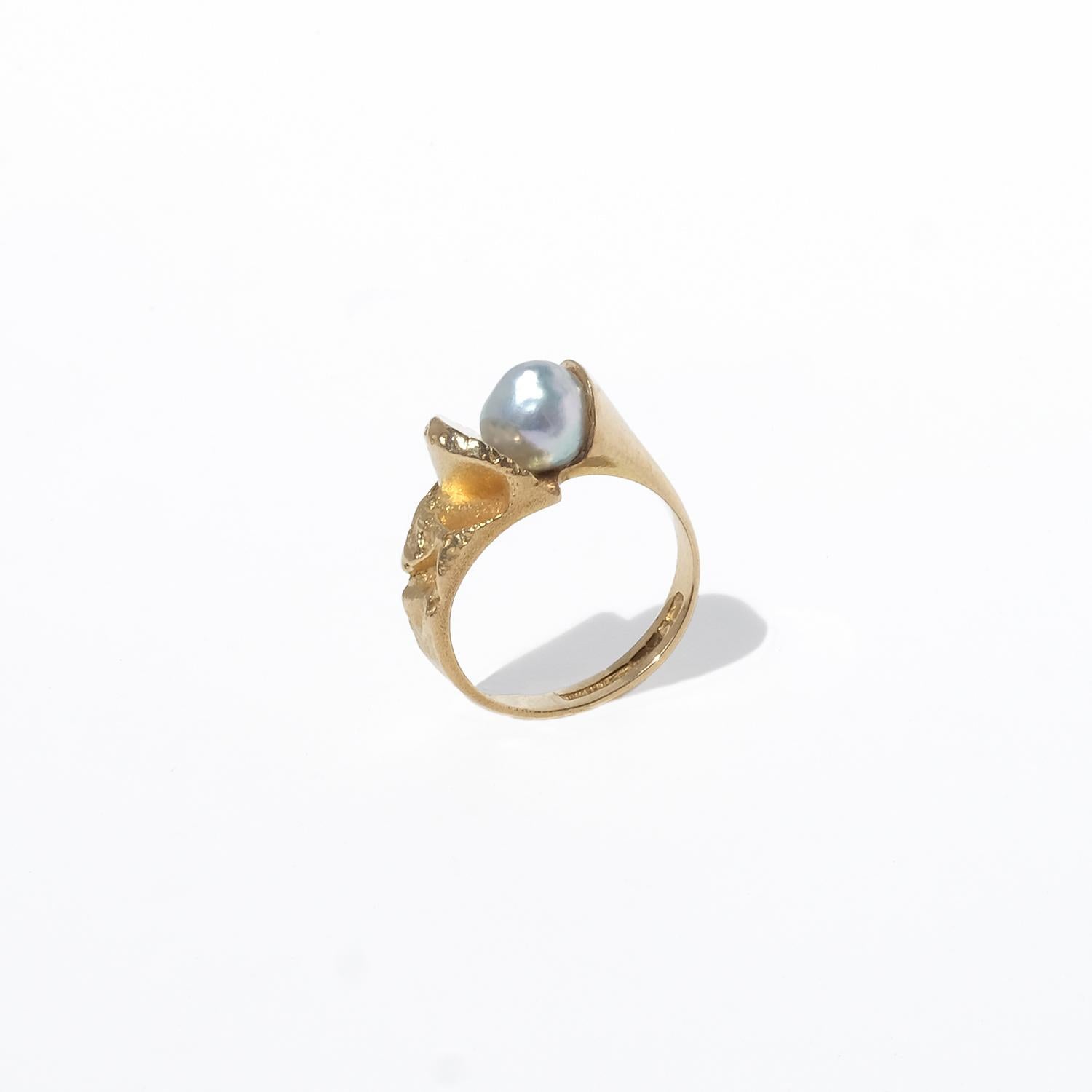 This unique ring, called Birth of a Pearl, is crafted in 18-karat gold and features a single greyish pearl set at the center. The design is distinctly asymmetrical, with one side of the pearl framed by highly polished gold in a smooth, curved form