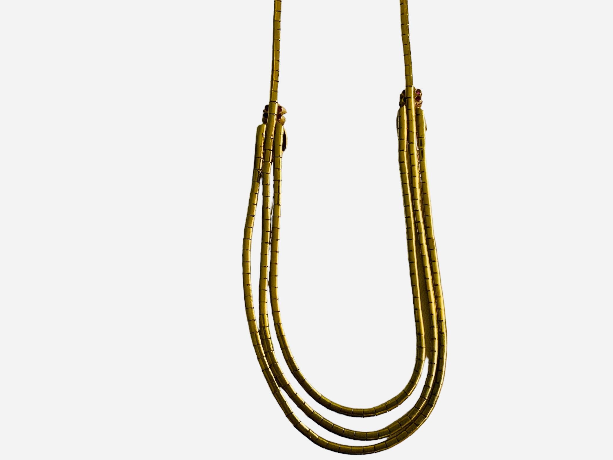 Questa è una collana in oro 18 carati e rubini. Raffigura tre catene in oro giallo composte da maglie a blocchi singoli unite da un fiore di rubini e ornate da un mazzo di foglie d'oro su ciascun lato per formare una collana a catena singola