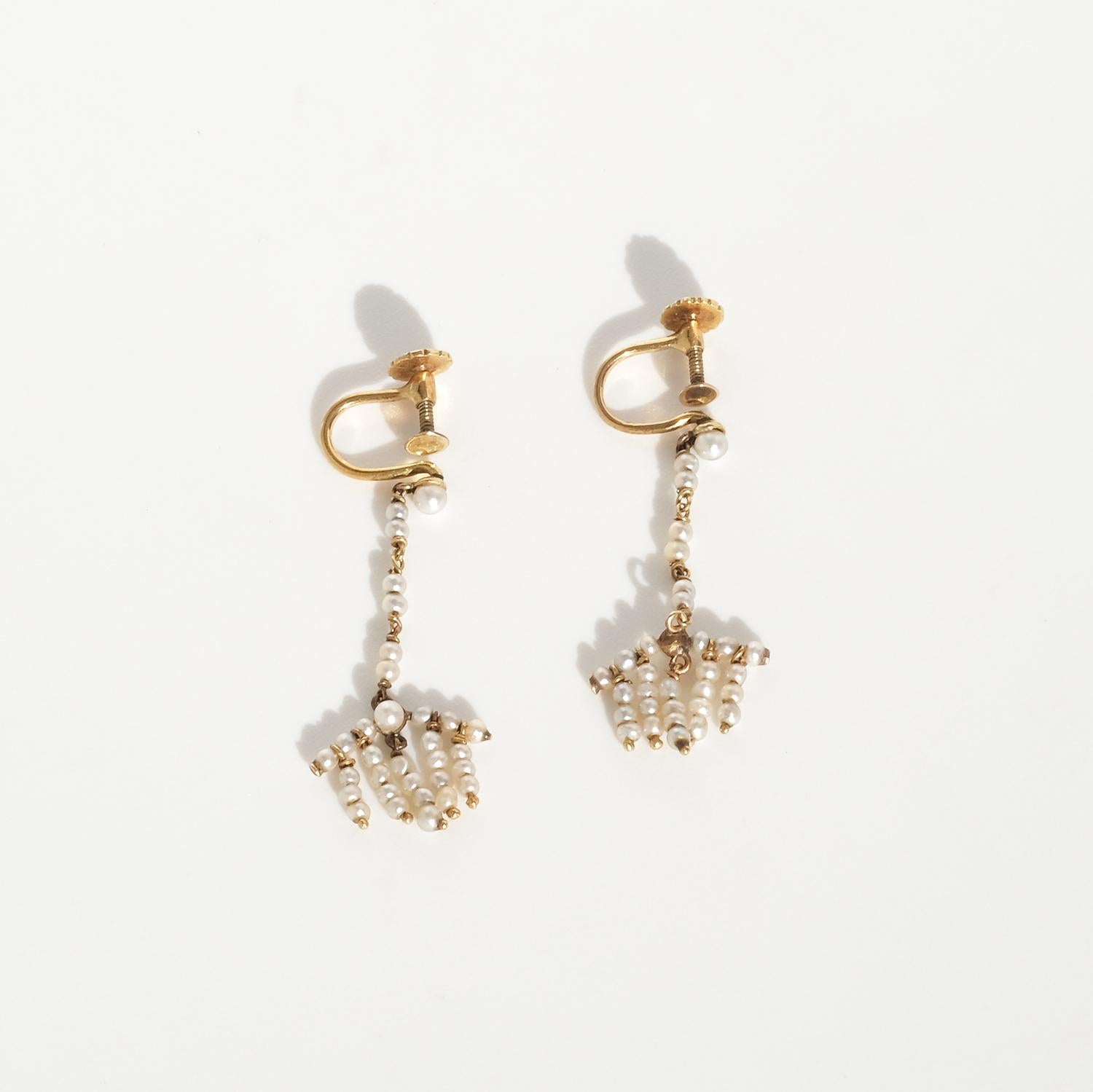 These 18 karat gold earrings feature delicate seed pearls arranged in cascading clusters, creating a chandelier-like effect. The design includes a slender vertical drop, allowing the earrings to hang elegantly and sway gently with movement. They are