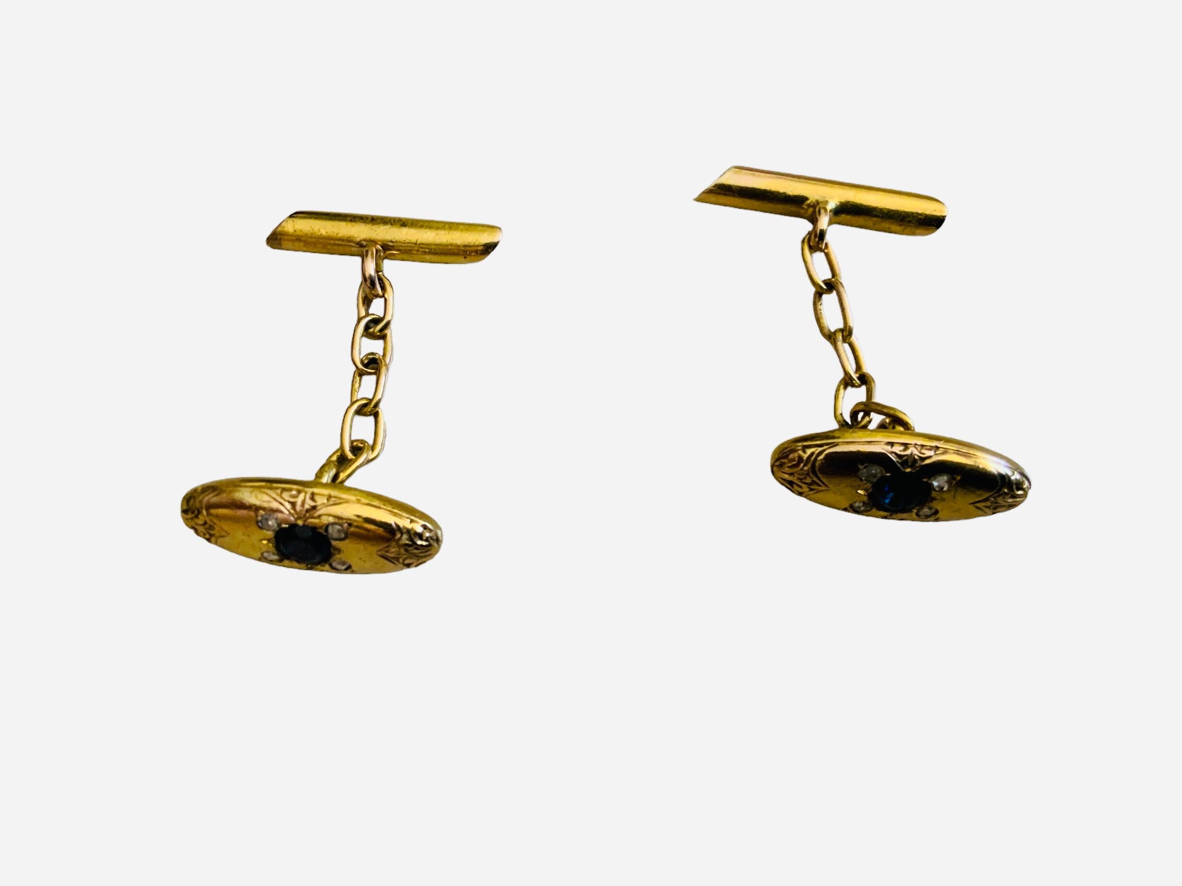 This is a 18K gold chain link pair of cufflinks. It depicts an oval shaped cufflinks engraved with some Fleur de Lis in each side. It is embellished in the center with a round blue topaz surrounded by one pave diamond, one pave sapphire and clear